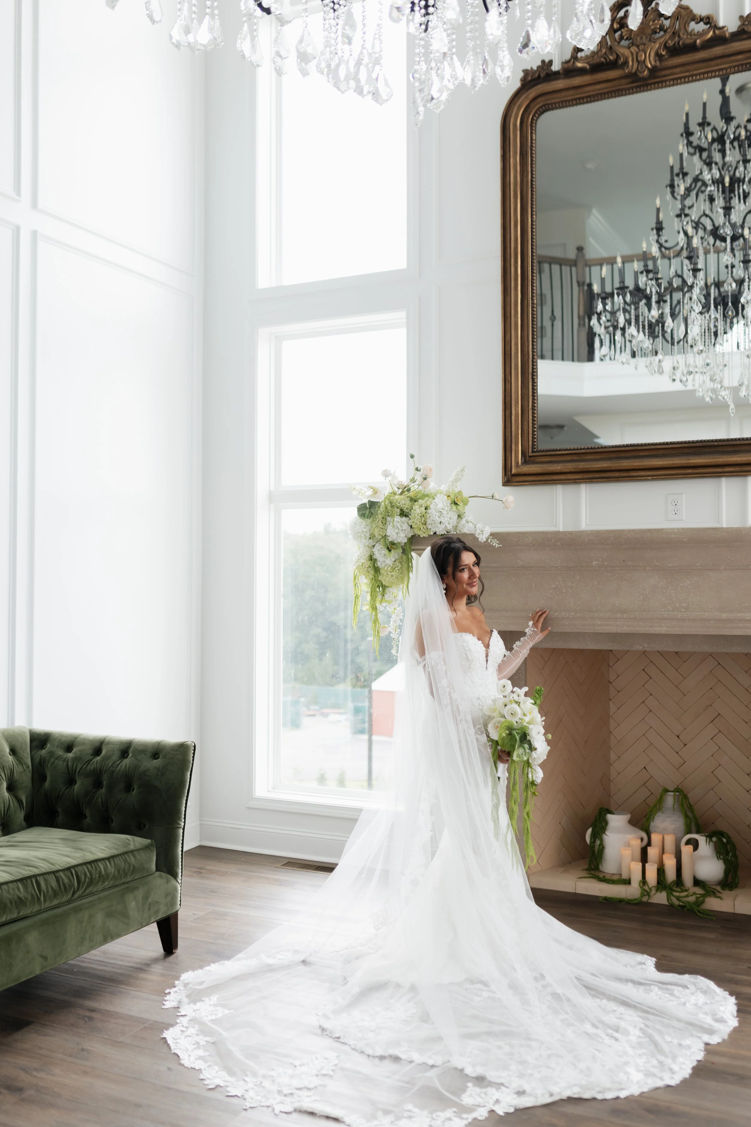 A bride in a white wedding dress and veil standing indoors next to a large mirror and a fireplace decorated with candles and greenery. She holds a bouquet of white flowers and greenery.