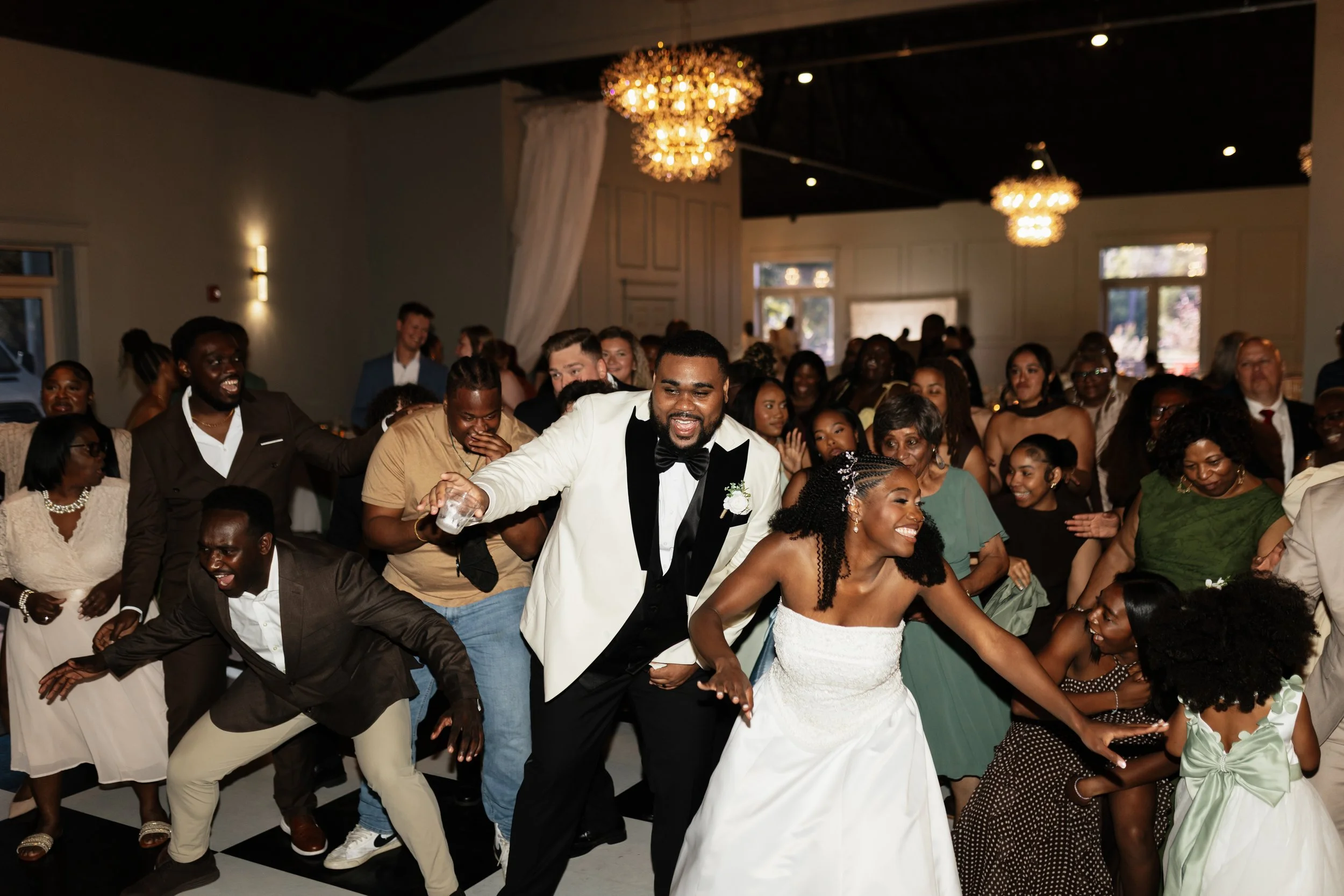 People dancing and celebrating at a wedding reception in a decorated indoor venue with chandeliers.