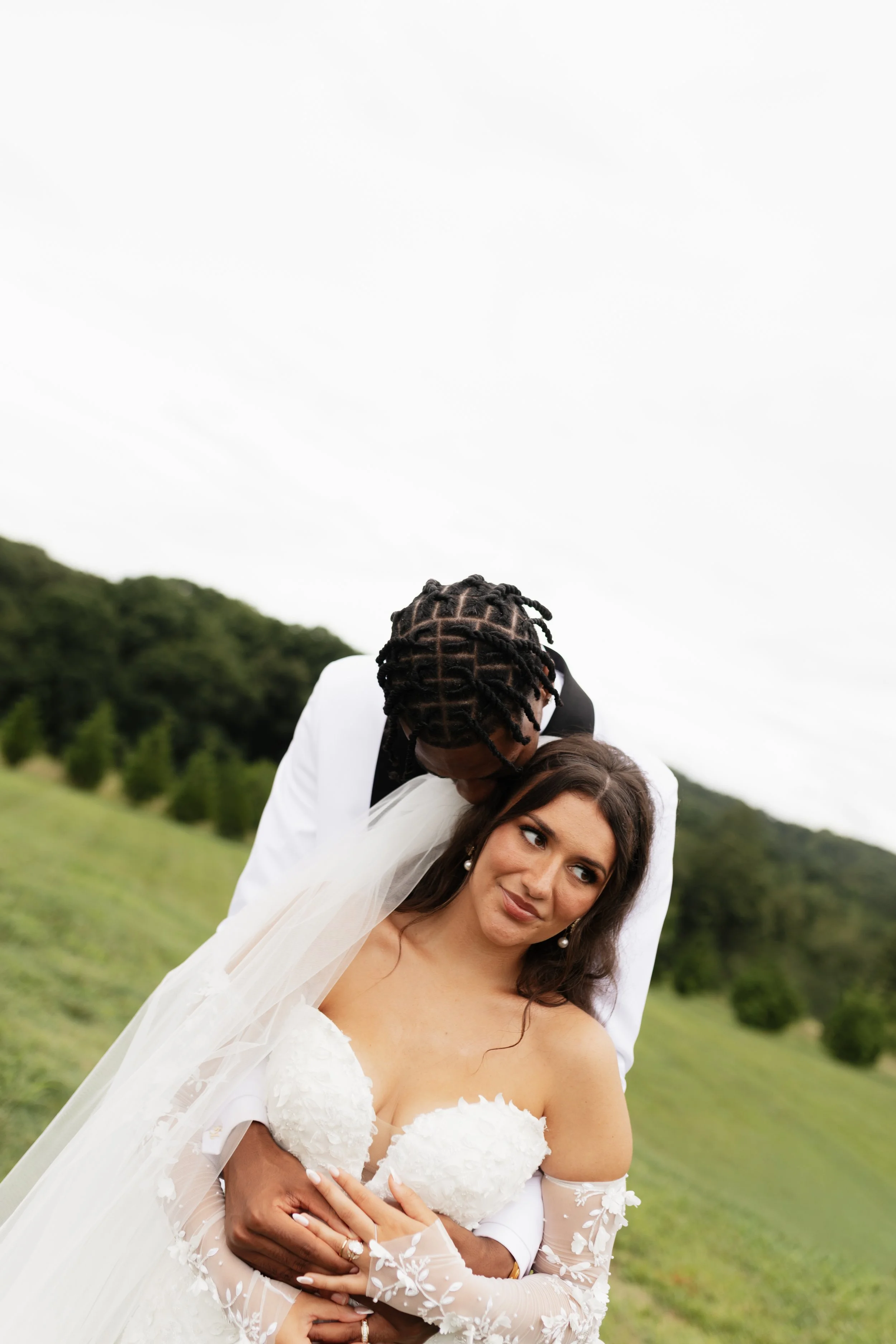 A bride in a white wedding gown and veil stands outdoors, while a groom with braided hair in a white suit gently kisses her head against a green hillside and cloudy sky background.