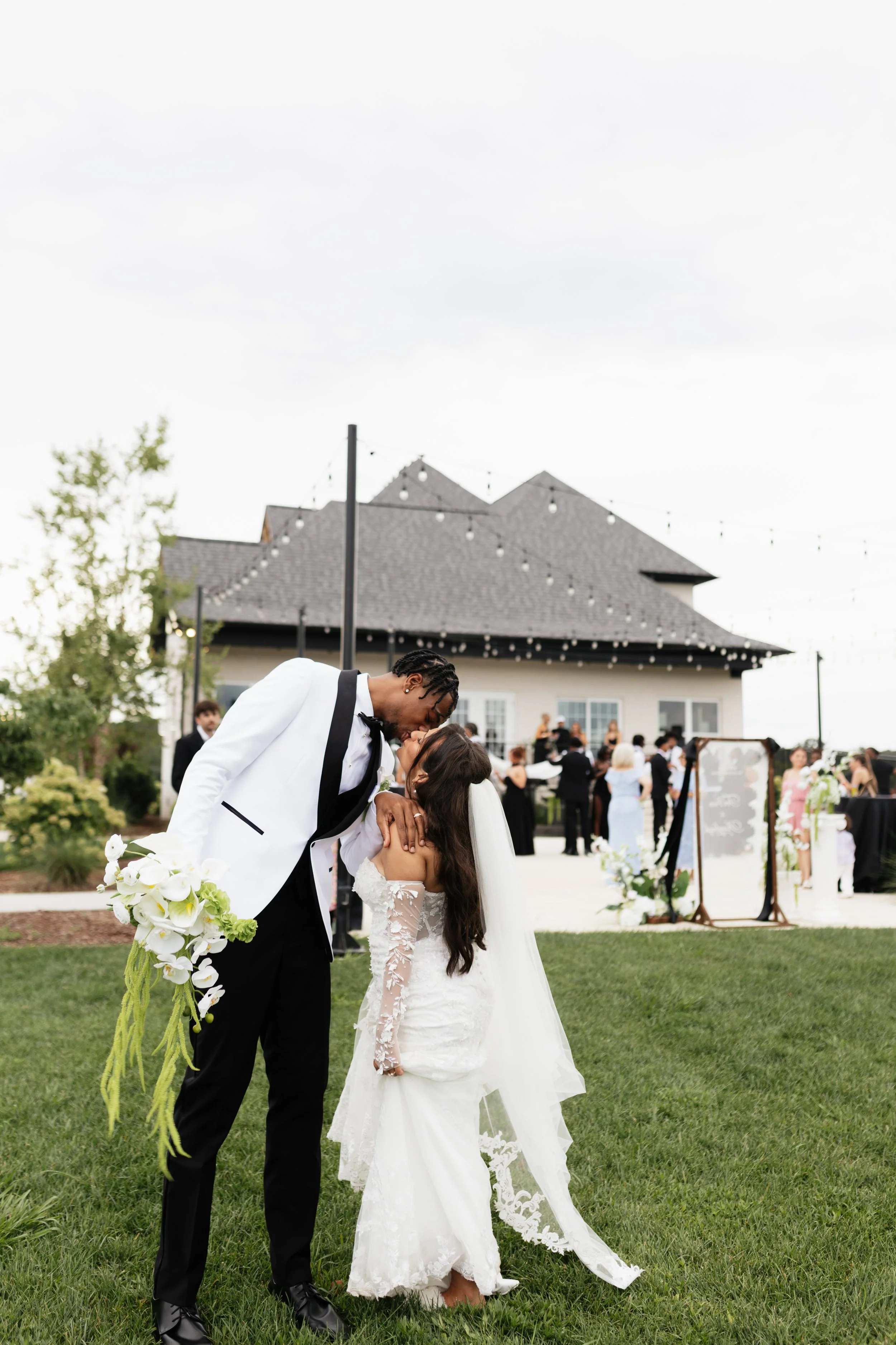 A couple sharing a kiss at an outdoor wedding ceremony, with the groom in a white tuxedo jacket and the bride in a lace wedding gown. The background shows a gathering of guests, a ballroom, and string lights hanging above.