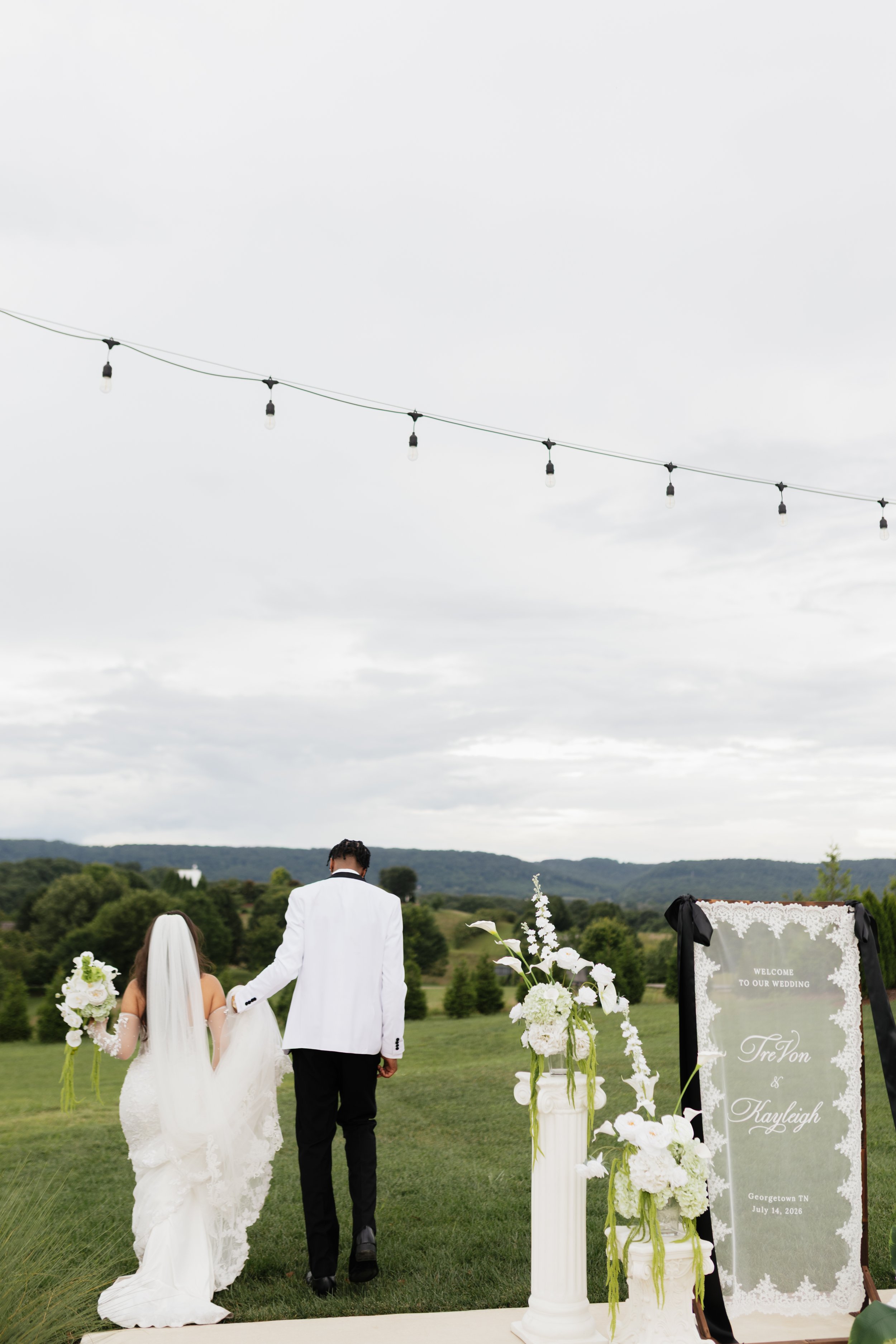 A bride and groom walking hand-in-hand outdoors at a wedding ceremony, with floral arrangements and a welcome sign on the right, overlooking a scenic landscape with trees and hills.