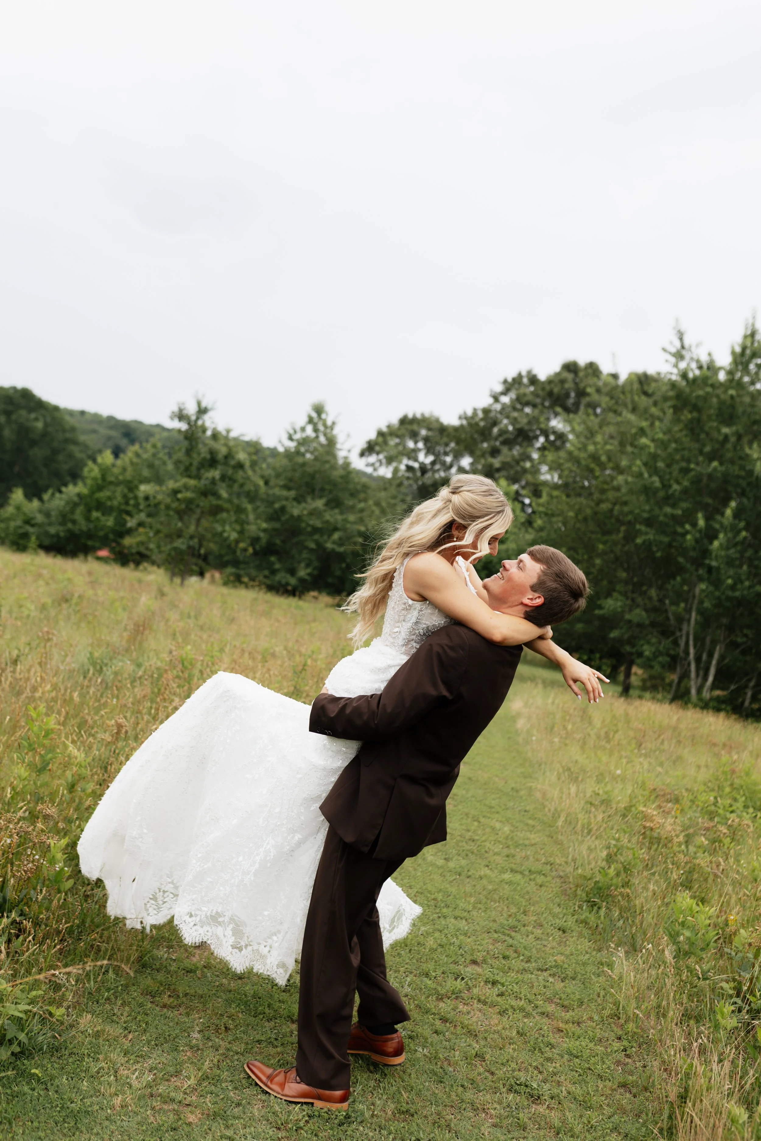 A man in a brown suit lifts a woman in a white wedding dress in a field with green grass and trees, under an overcast sky.