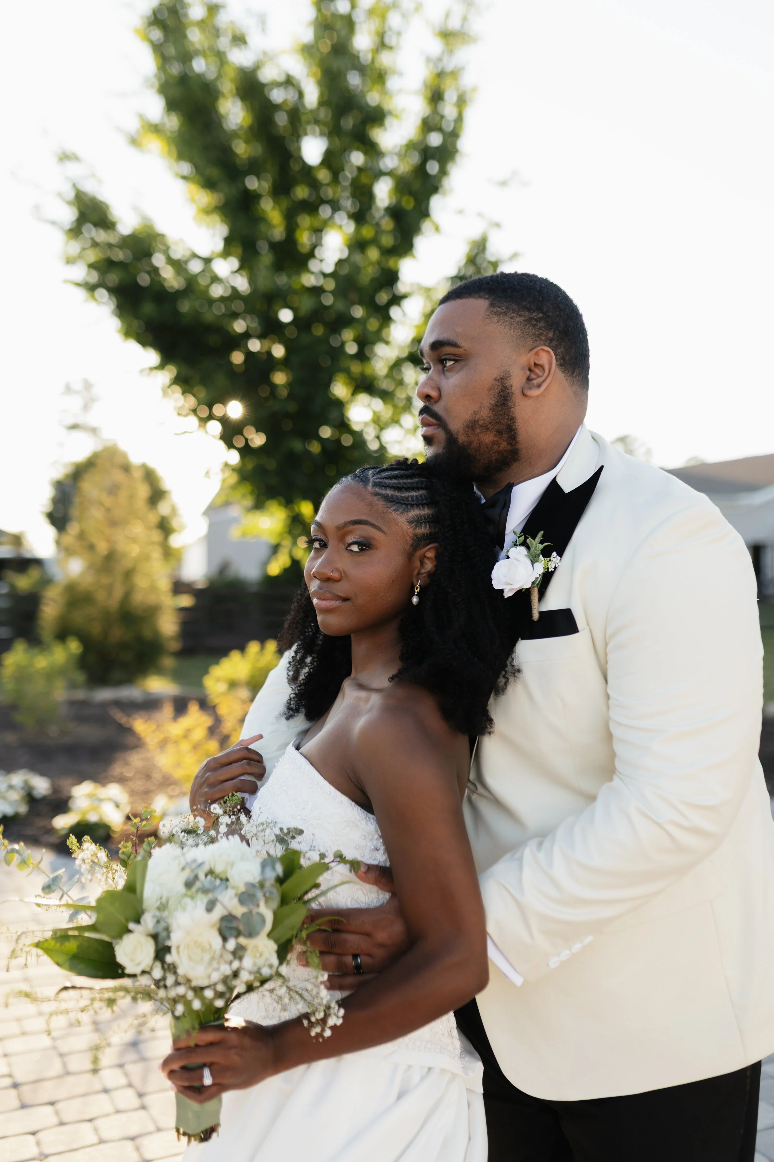 A bride and groom posing outdoors on their wedding day, with the bride holding a bouquet of white flowers.