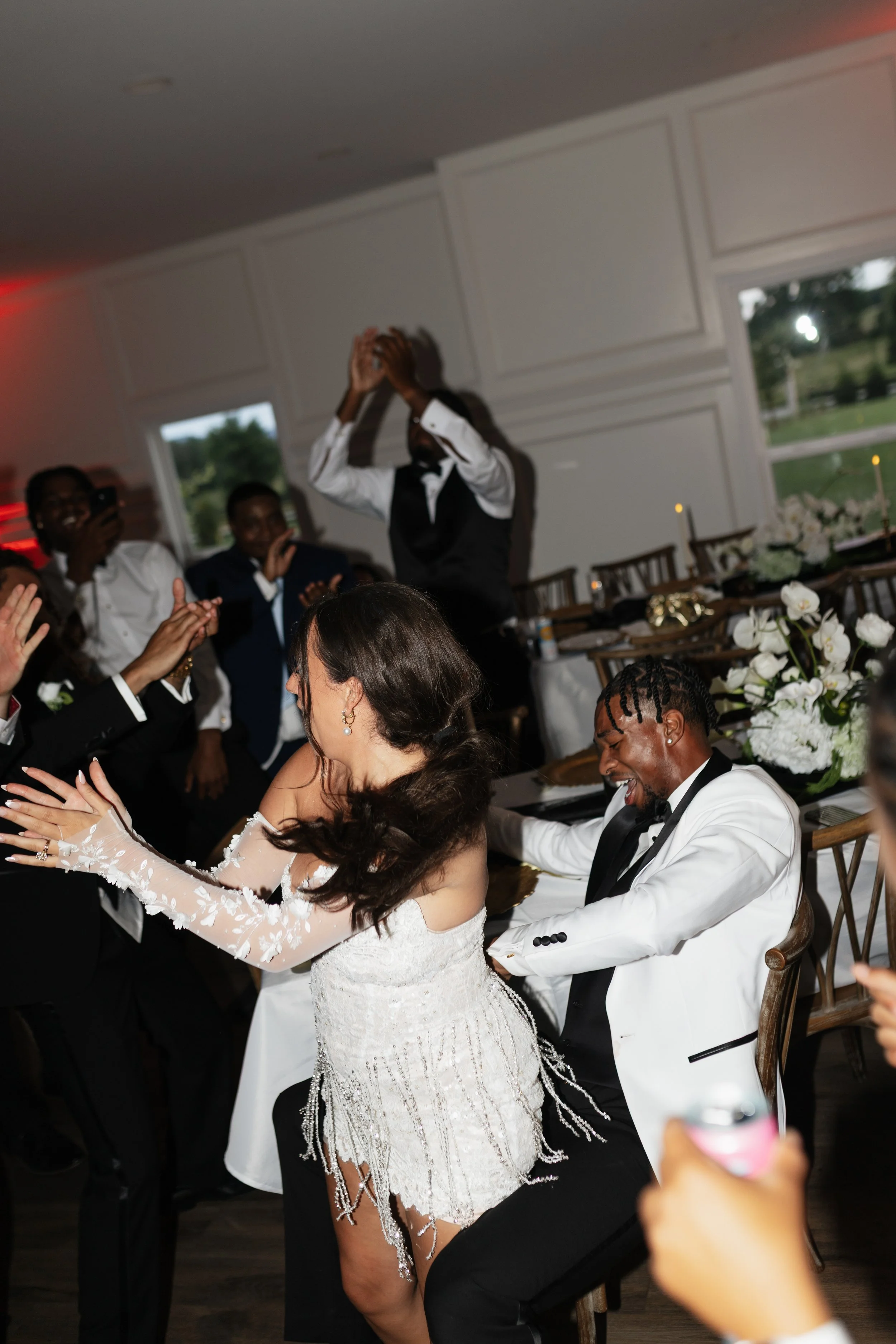 A wedding celebration indoors with a bride, groom, and guests dancing and smiling, decorated table with white flowers, and large windows showing a green outdoor landscape.