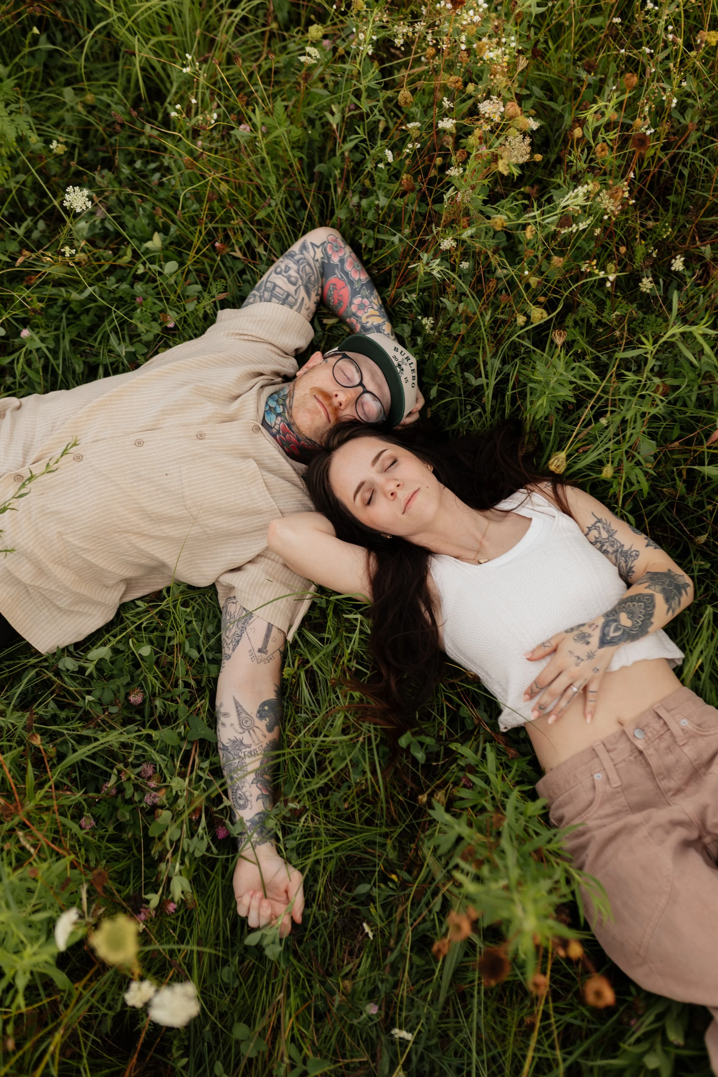 A man and woman lying on their backs in a field of tall grass and wildflowers with their eyes closed, appearing relaxed and peaceful.