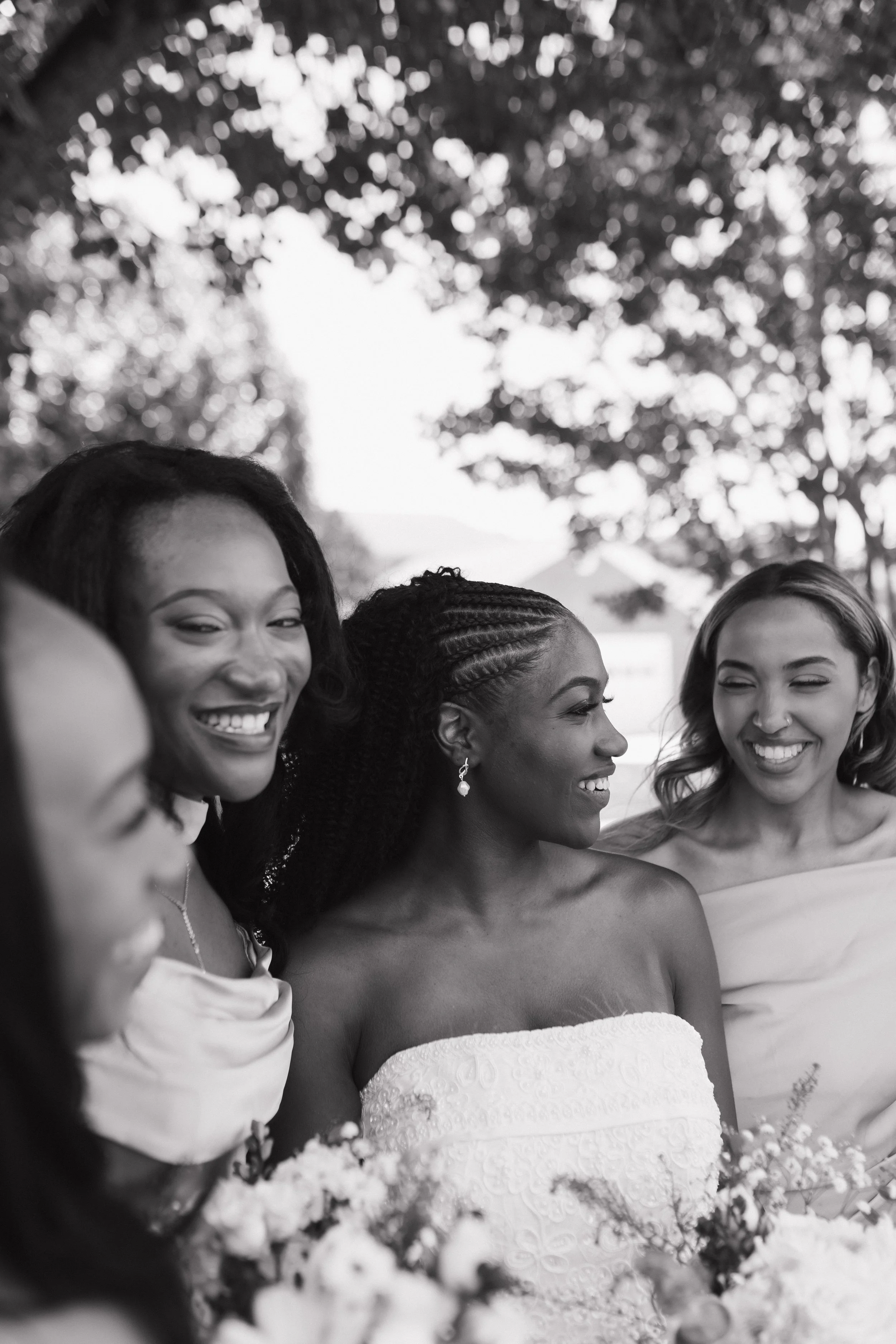 Black and white photo of five women smiling and looking at each other, dressed in strapless dresses, outdoors with trees in the background.