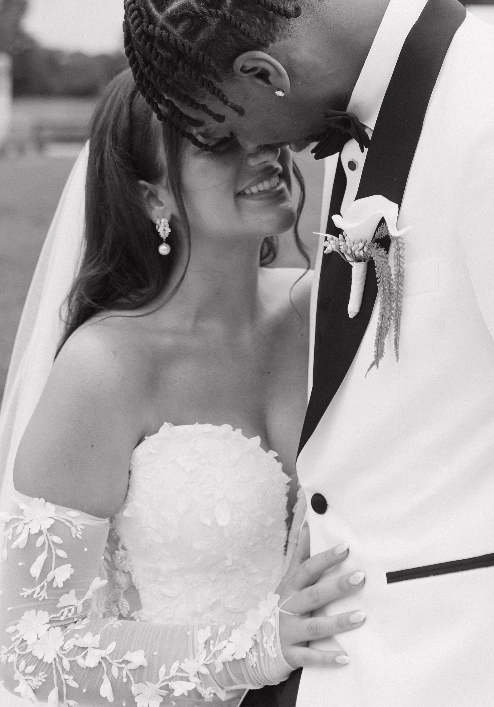 A bride and groom share a tender moment, with the groom bending down to gently touch his forehead to the bride's head, both smiling softly. The bride is wearing a wedding dress with floral lace details and pearl earrings, while the groom is dressed i