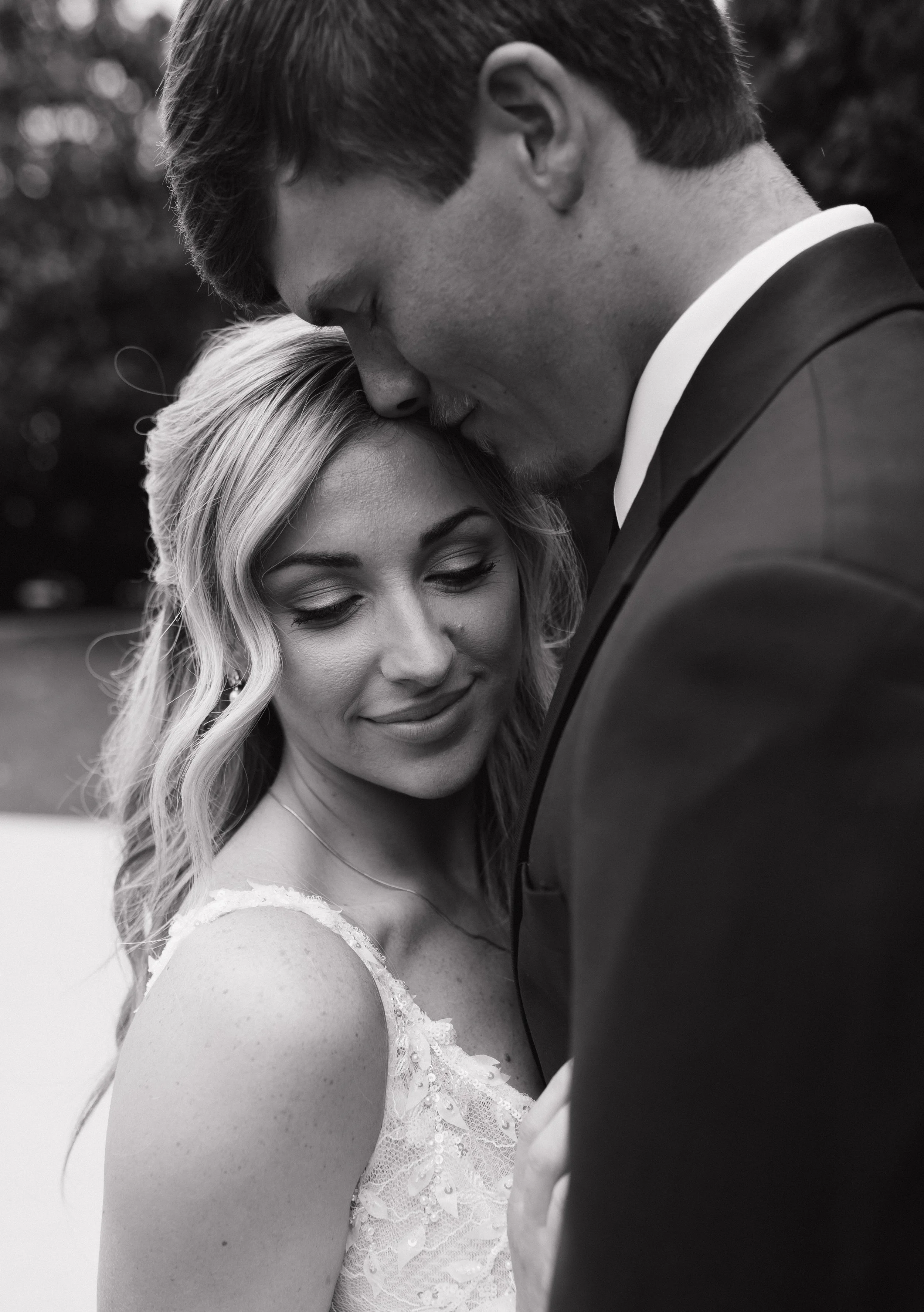 A black and white close-up photo of a couple on their wedding day, with the man gently touching his forehead to the woman's head, both with their eyes closed.