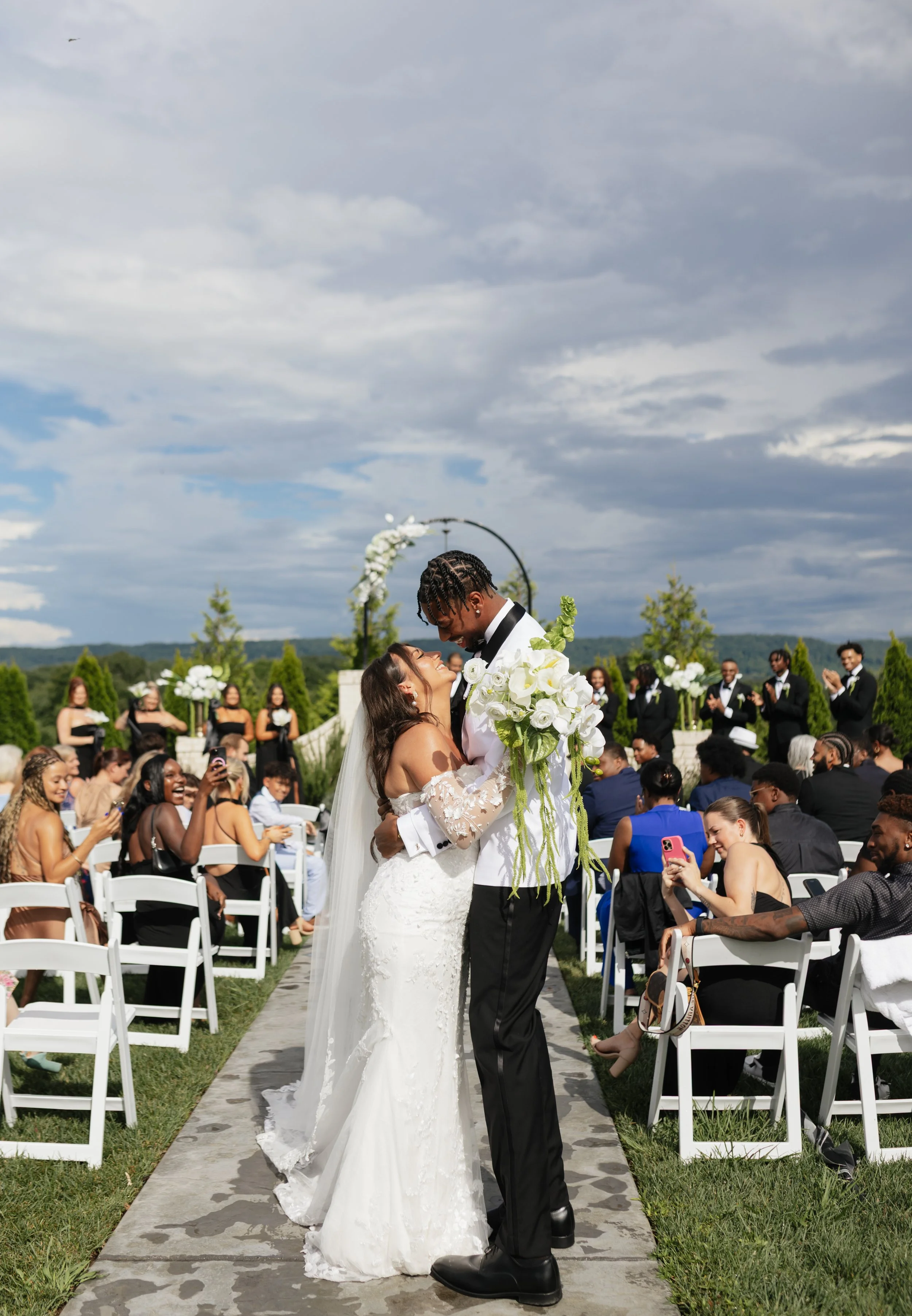A wedding ceremony outdoors with the bride and groom standing on a stone aisle, holding hands, surrounded by seated guests and bridesmaids and groomsmen. The sky is partly cloudy.