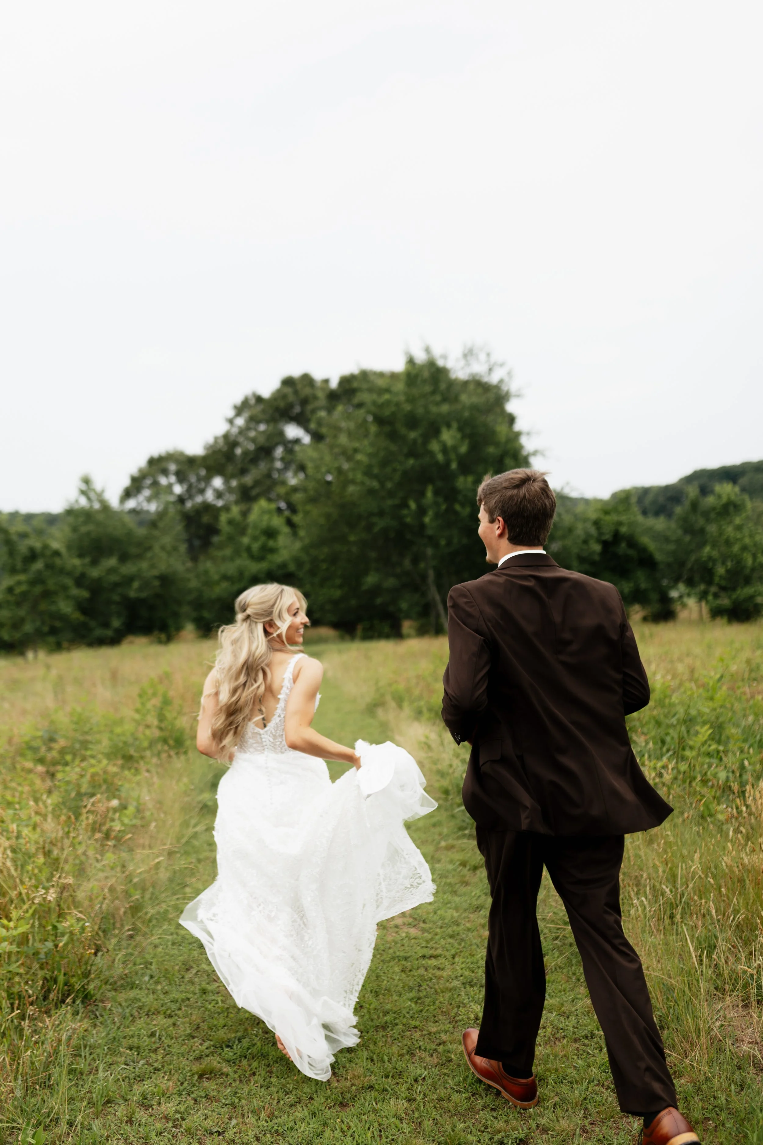 A bride and groom walking through a grassy field with trees in the background on a cloudy day.