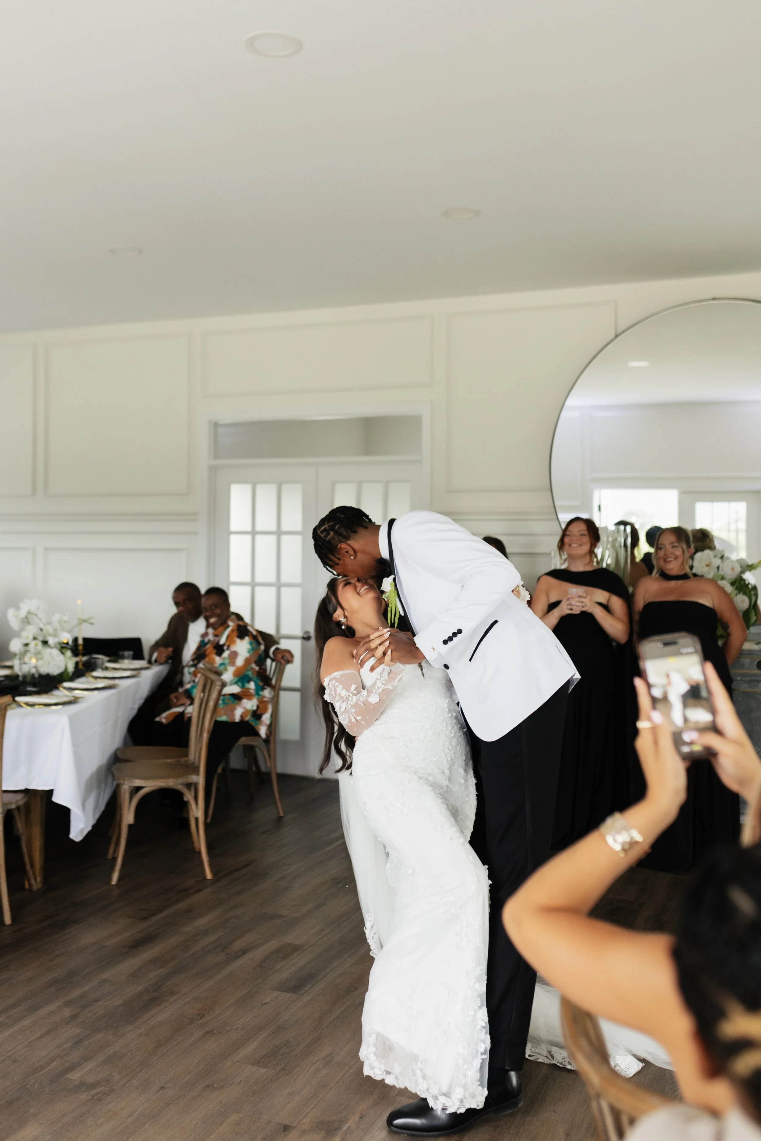 A bride and groom dance at their wedding reception, with guests watching and smiling in a bright, elegant room.