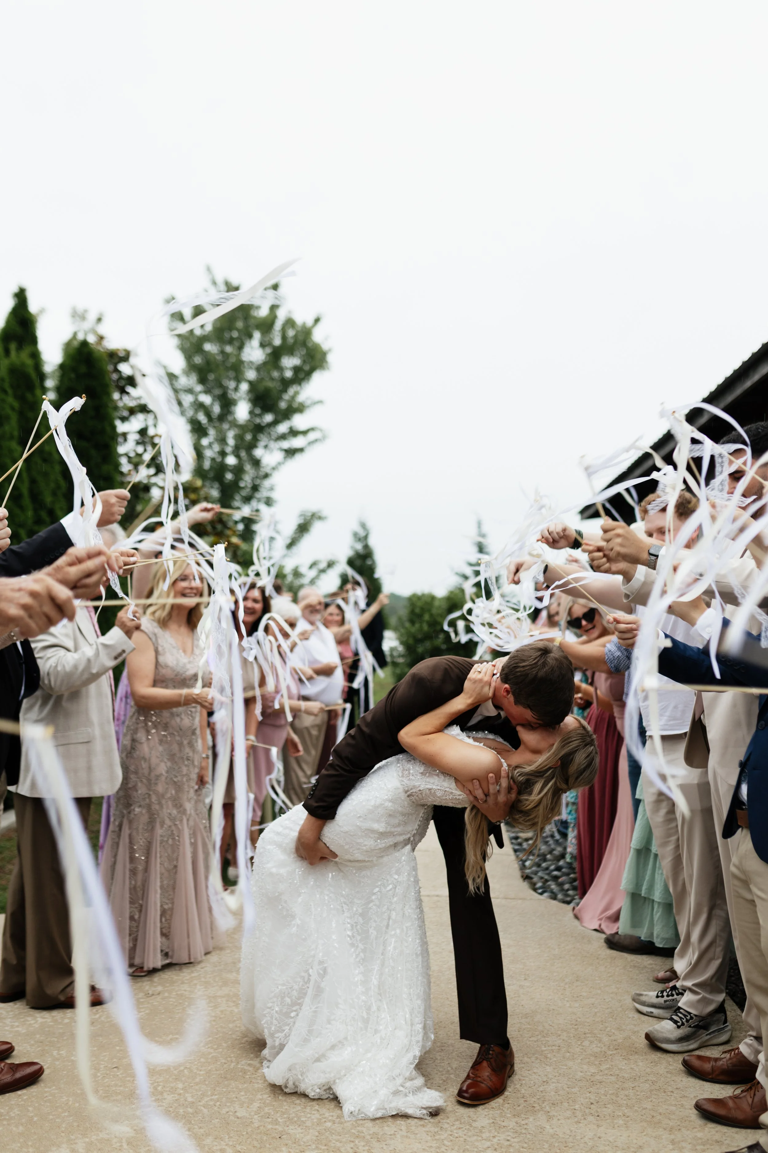 A newlywed couple sharing a kiss in front of friends and family at their outdoor wedding reception, with guests holding white streamers in celebration.