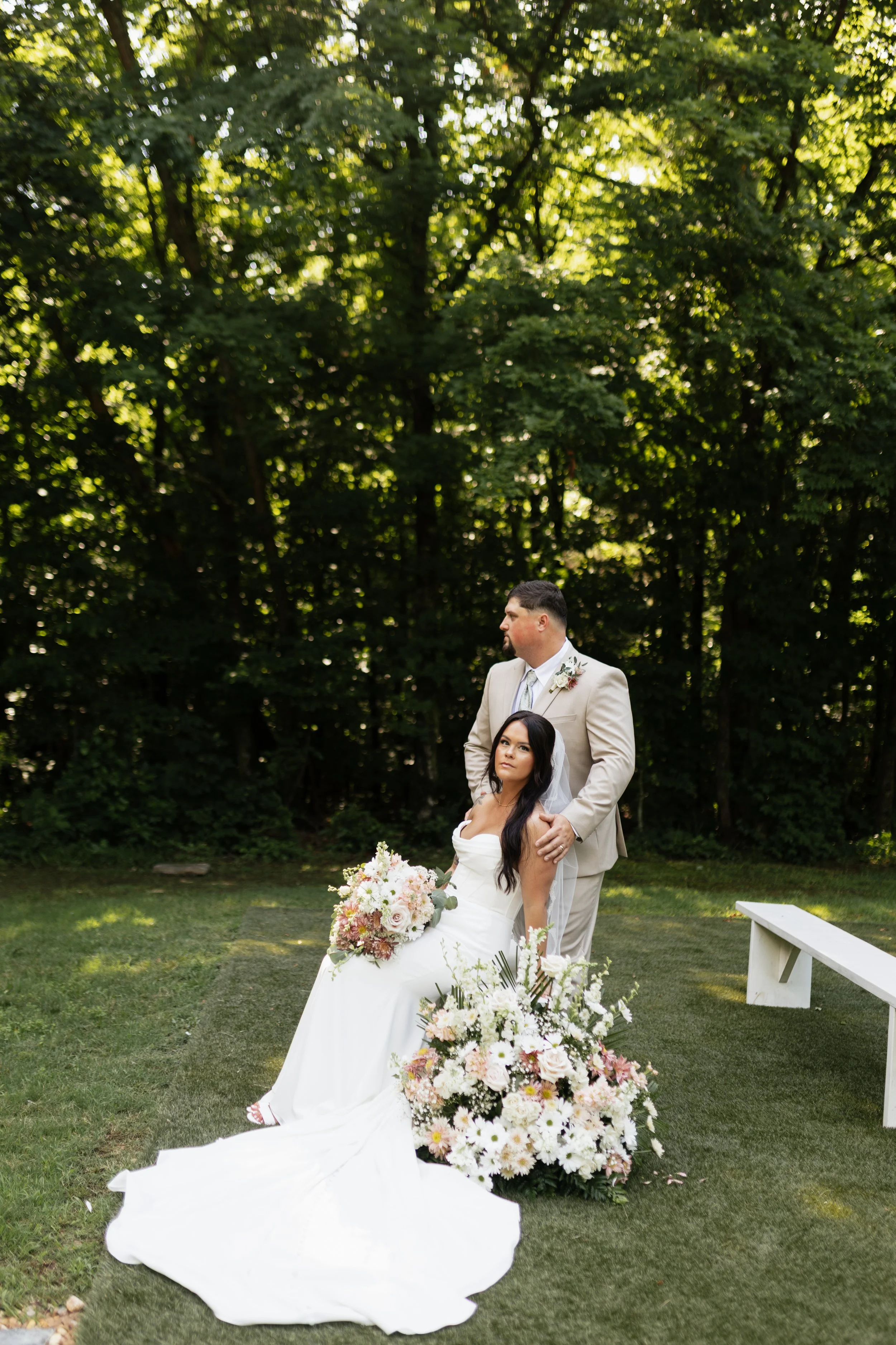 A bride and groom standing outdoors in a garden with green trees in the background, during a wedding ceremony. The bride is sitting on a white bench, holding a large bouquet of white and blush flowers, while the groom stands behind her with his hands