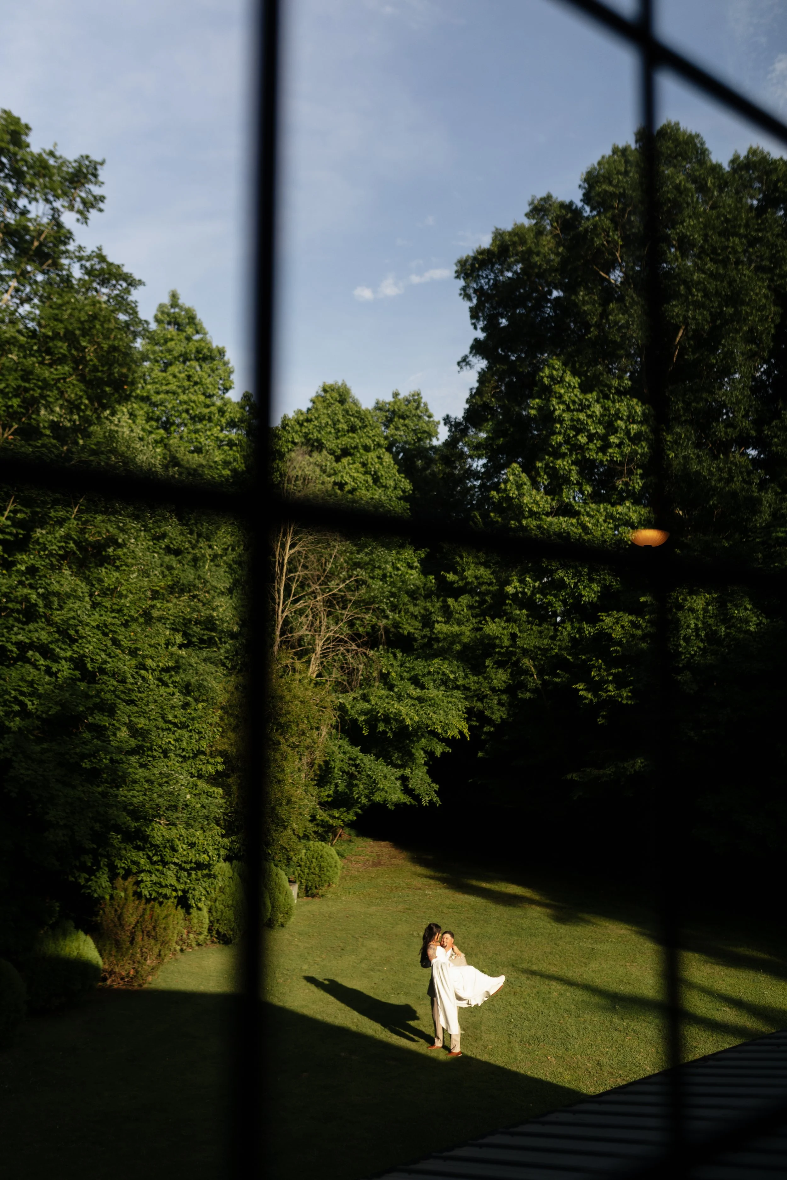 A couple dressed in white, with the man holding the woman in his arms, standing on a grassy yard surrounded by trees, seen through a dark fence with vertical bars, during the daytime.