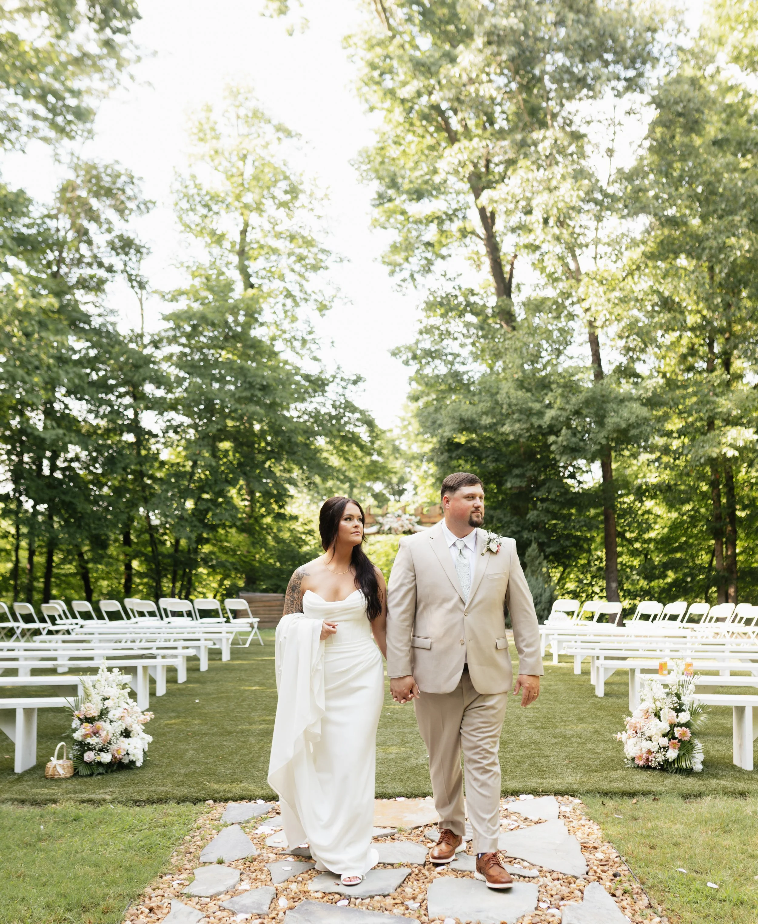 A bride and groom walking hand in hand down the aisle outdoors during a wedding ceremony, with white chairs and floral arrangements in the background and lush green trees overhead.