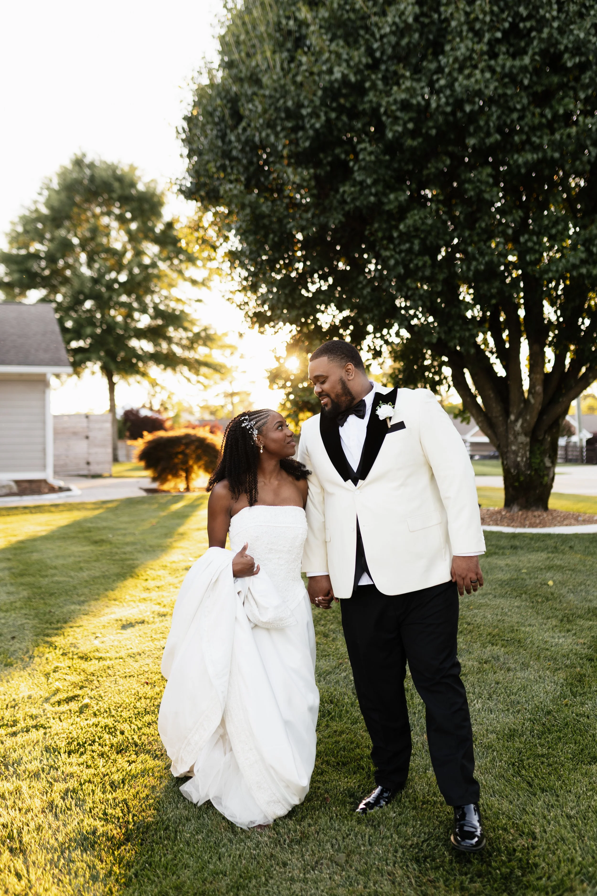 A bride and groom walk hand-in-hand outdoors in a park during sunset, with large trees and a house in the background, smiling at each other.