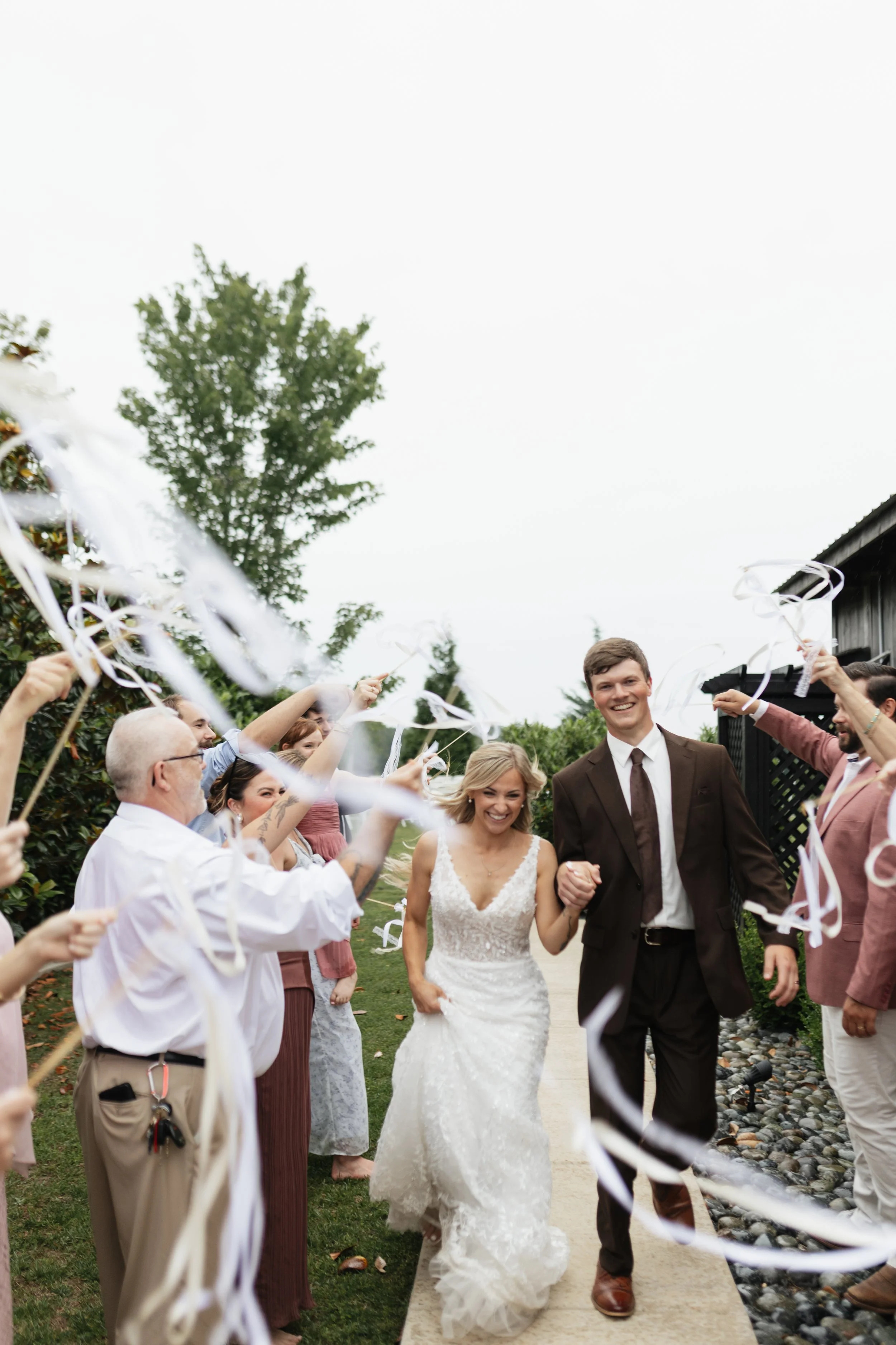 A bride and groom walking down an outdoor aisle while guests celebrate and throw streamers. The bride wears a white lace wedding gown, and the groom wears a dark suit with a tie. The scene is lively with a joyful mood.