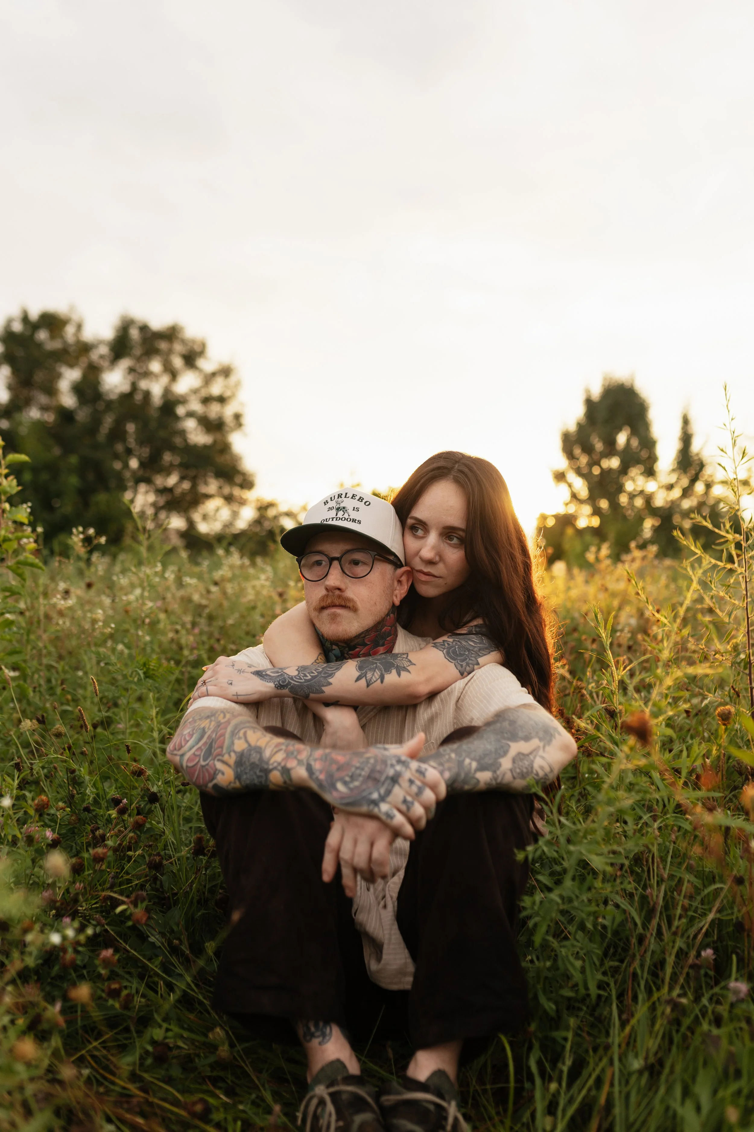 A man with tattoos wearing glasses and a baseball cap is sitting in a field with plants and flowers, with a woman with long brown hair hugging him from behind. They are outdoors during sunset, with trees and a cloudy sky in the background.
