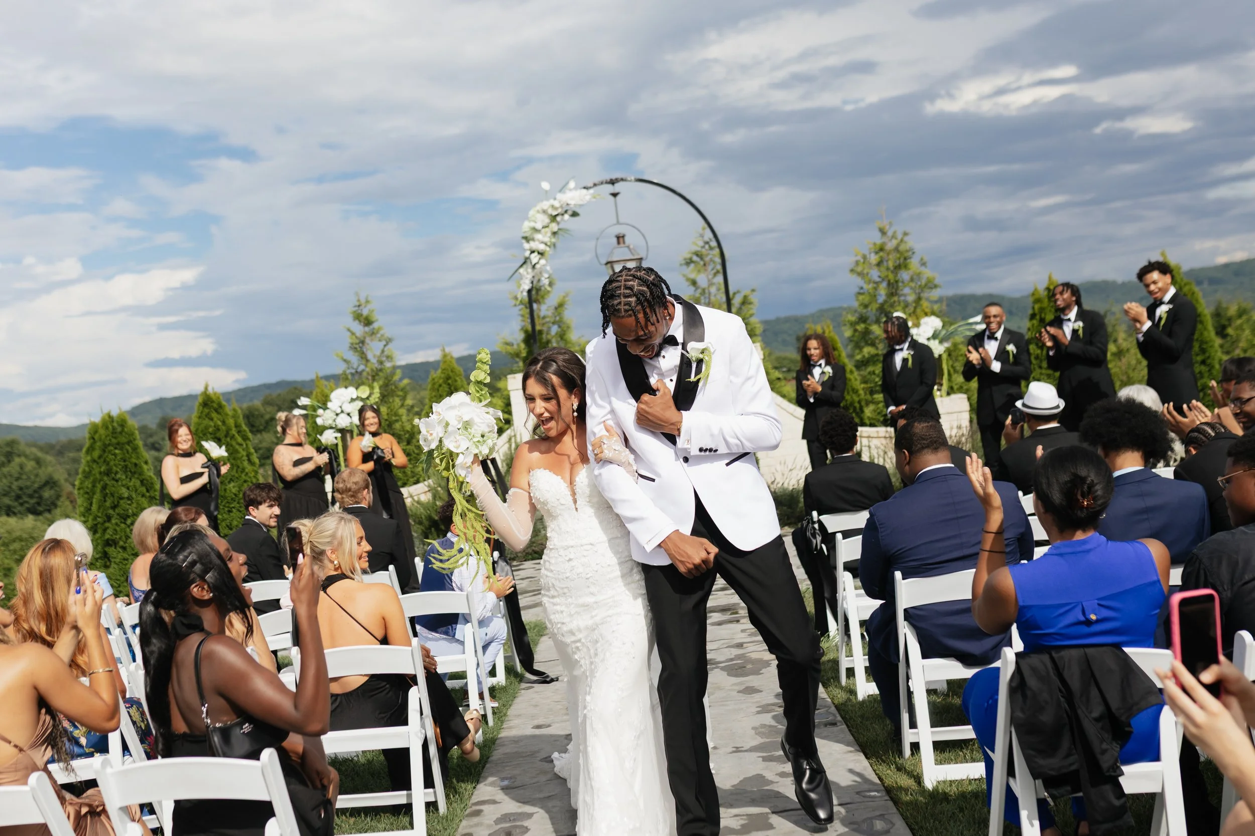 A couple, the groom in a white tuxedo with black lapel and the bride in a white lace wedding gown, dancing at an outdoor wedding ceremony with guests seated on white chairs and smiling in the background. The scene is set against a backdrop of hills a