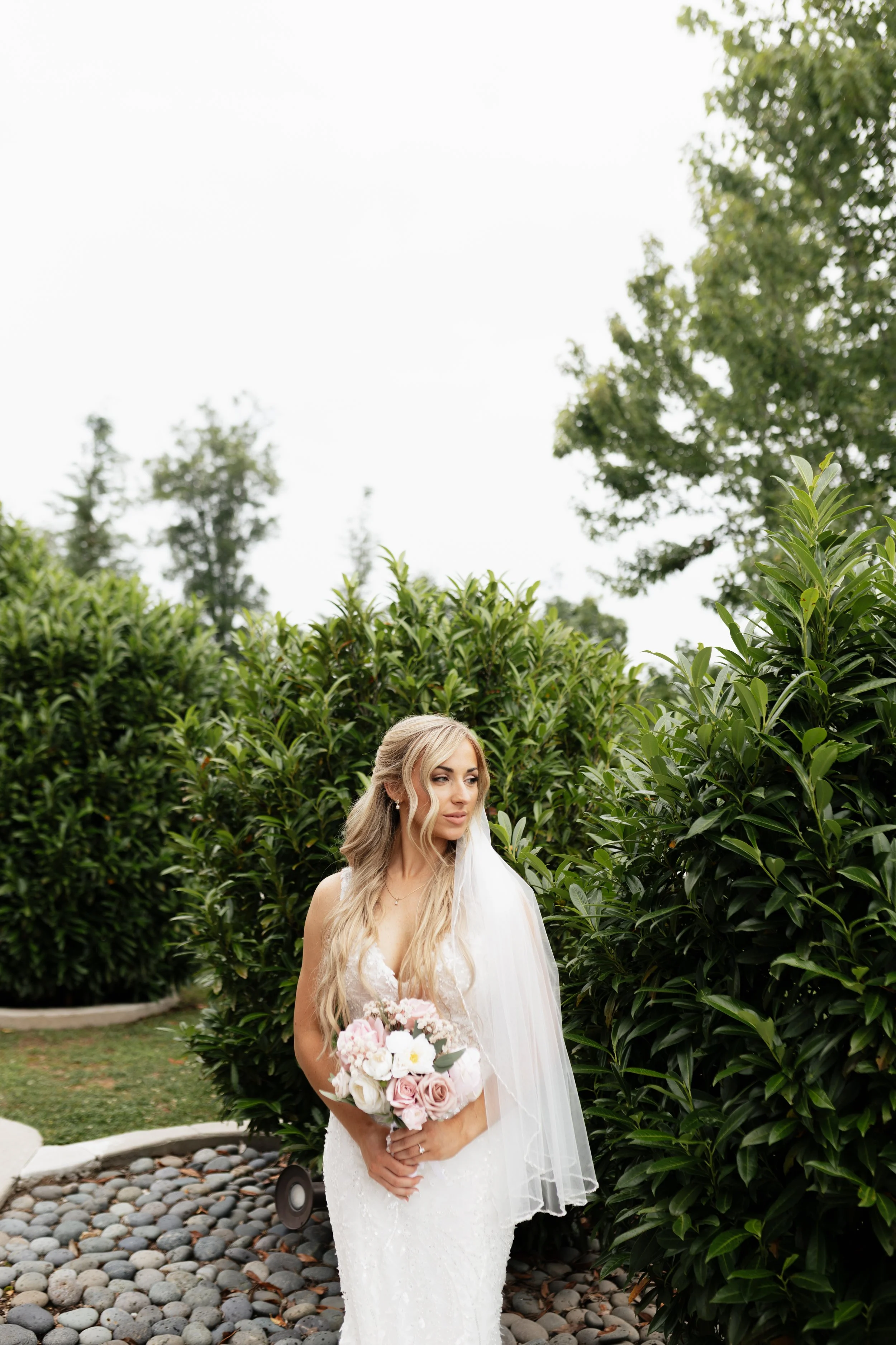 A bride with long blonde hair, wearing a white wedding dress with lace details and a veil, holding a bouquet of pink and white flowers, standing outdoors in front of green bushes and trees.