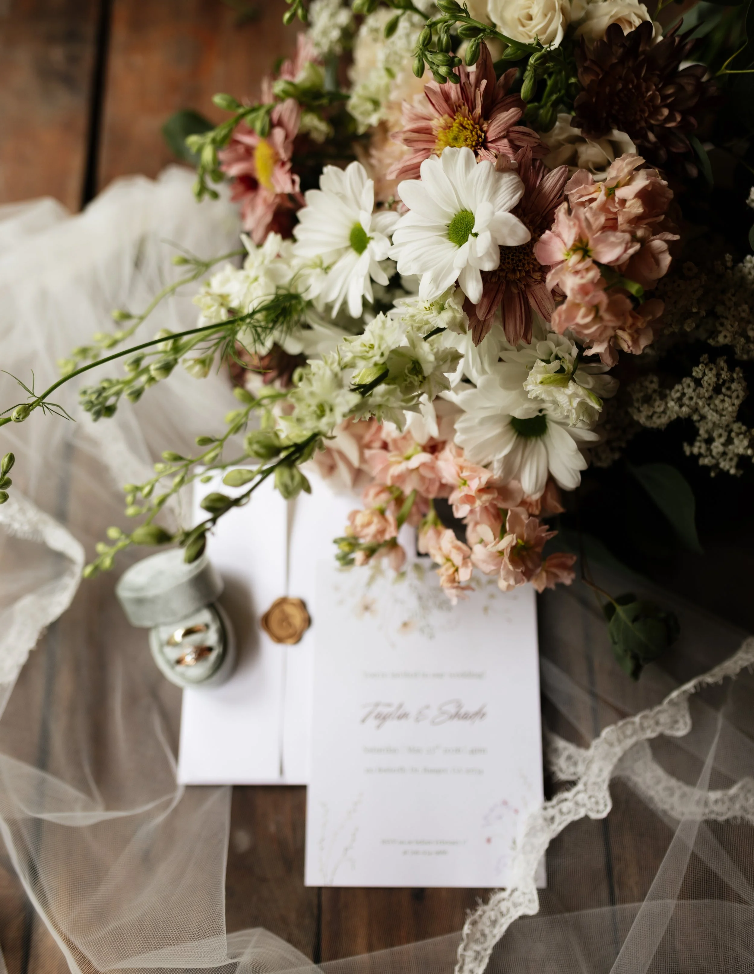 A wedding invitation card lying on a table with a bouquet of white, pink, and dark flowers, and a piece of lace fabric surrounding it.