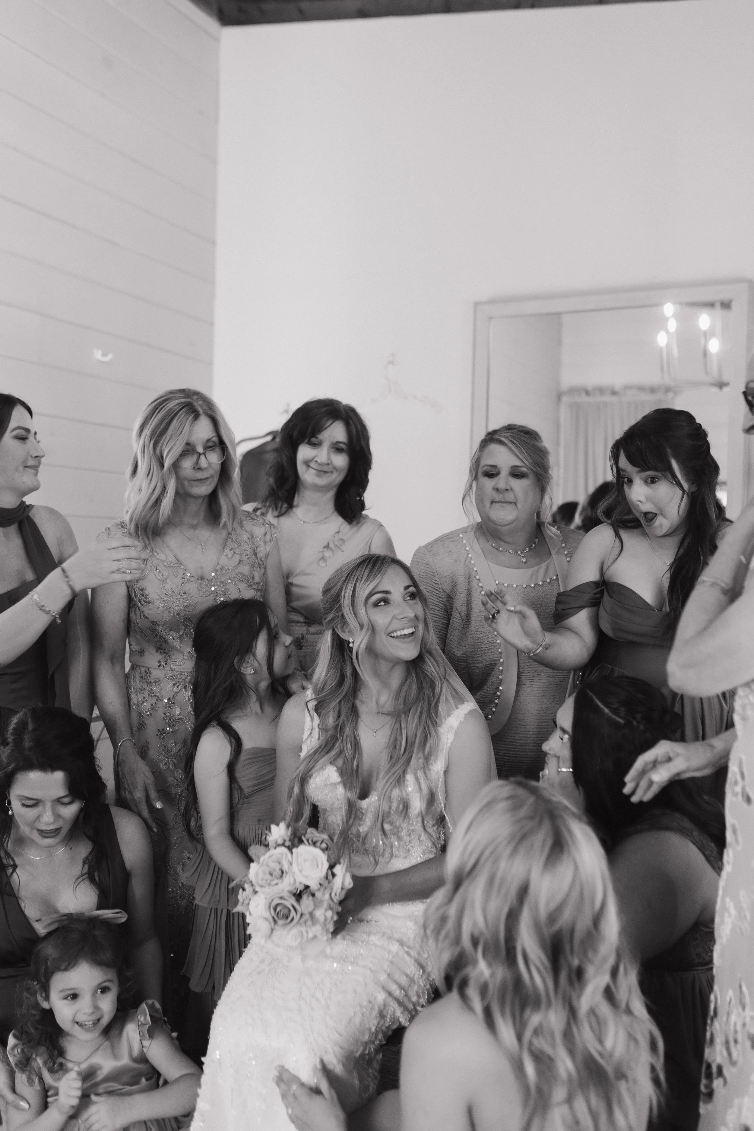 A group of women and children gathered around a bride holding a bouquet of flowers, at a wedding celebration in a decorated indoor setting.