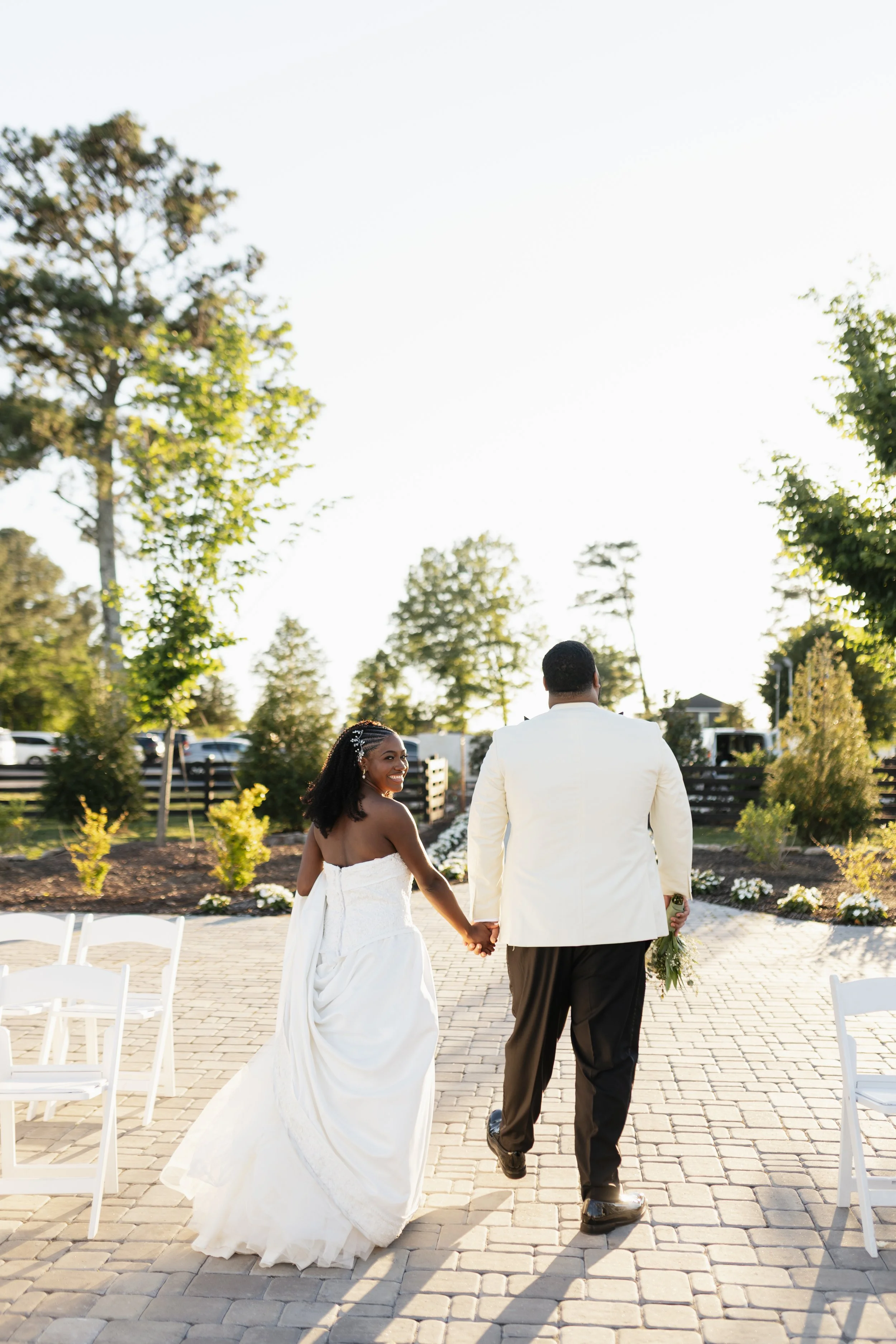 A bride and groom holding hands, walking away from the camera outdoors during daytime, surrounded by chairs, trees, and flowers, with a bright sky above.