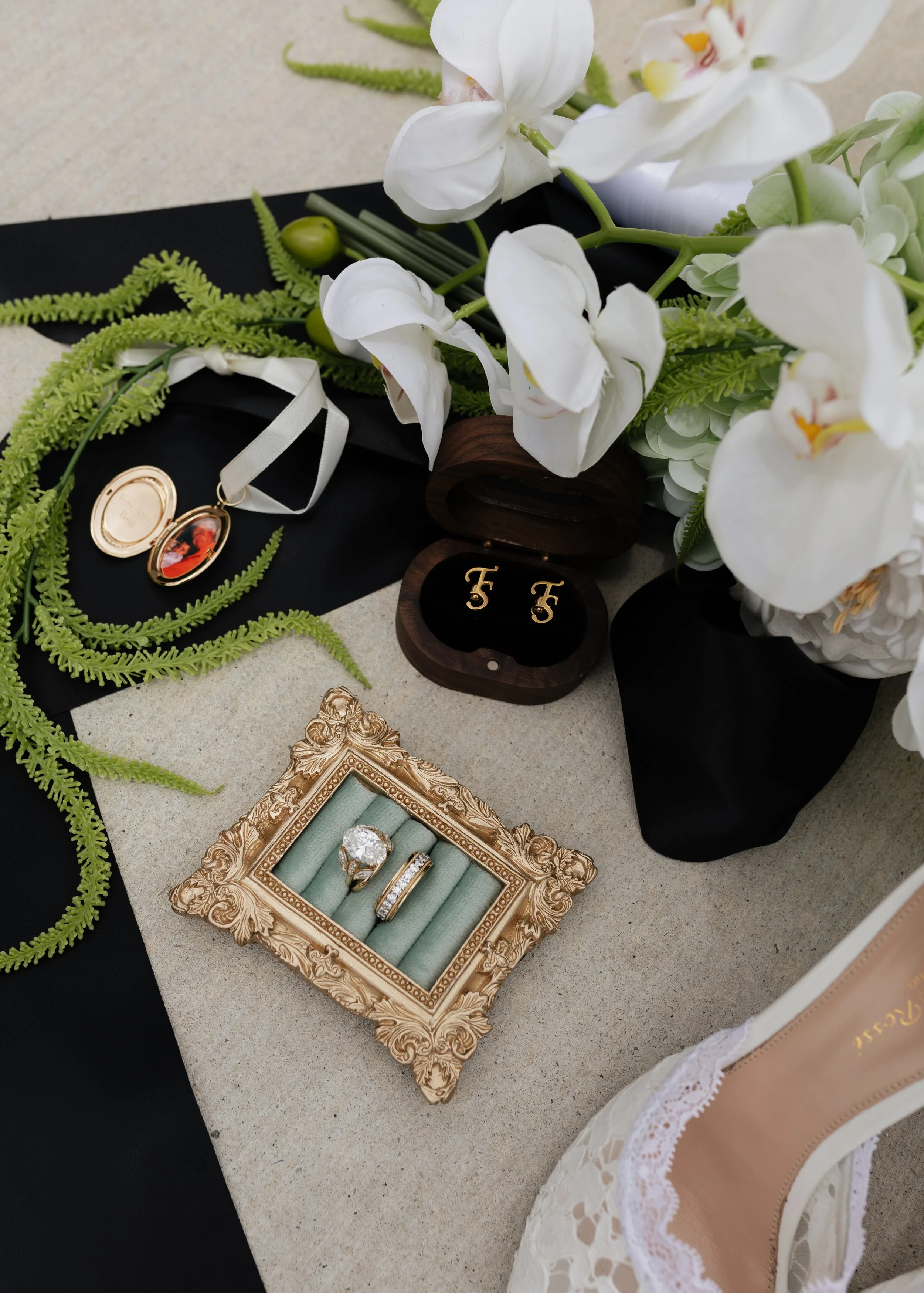 A floral arrangement of white orchids and green ferns, a jewelry box with diamond rings, and a pair of gold earrings with a black background and a beige border.