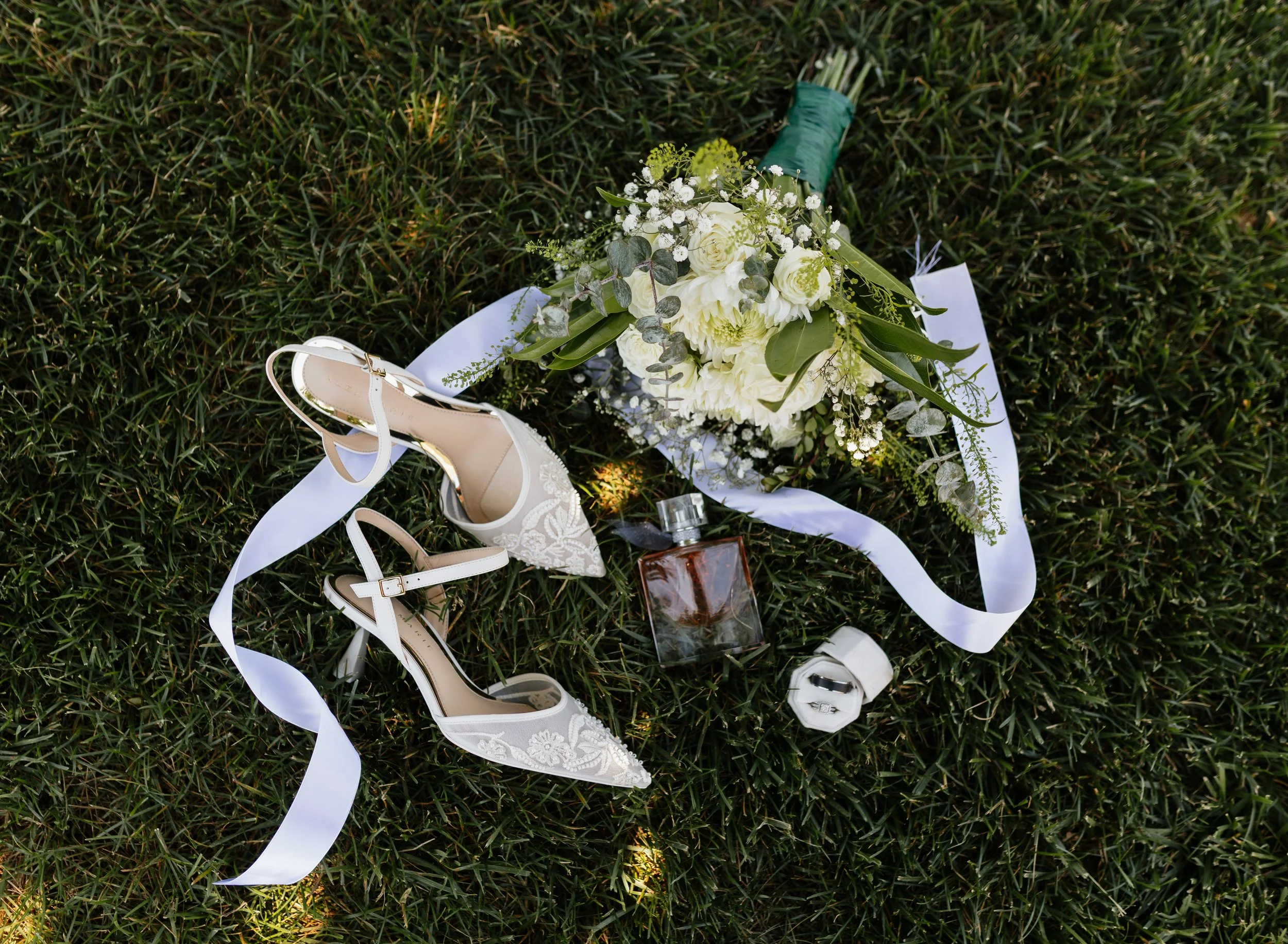 A bridal bouquet with white flowers and greenery lies on grass, next to white wedding shoes, a white ribbon, and a bottle of perfume.