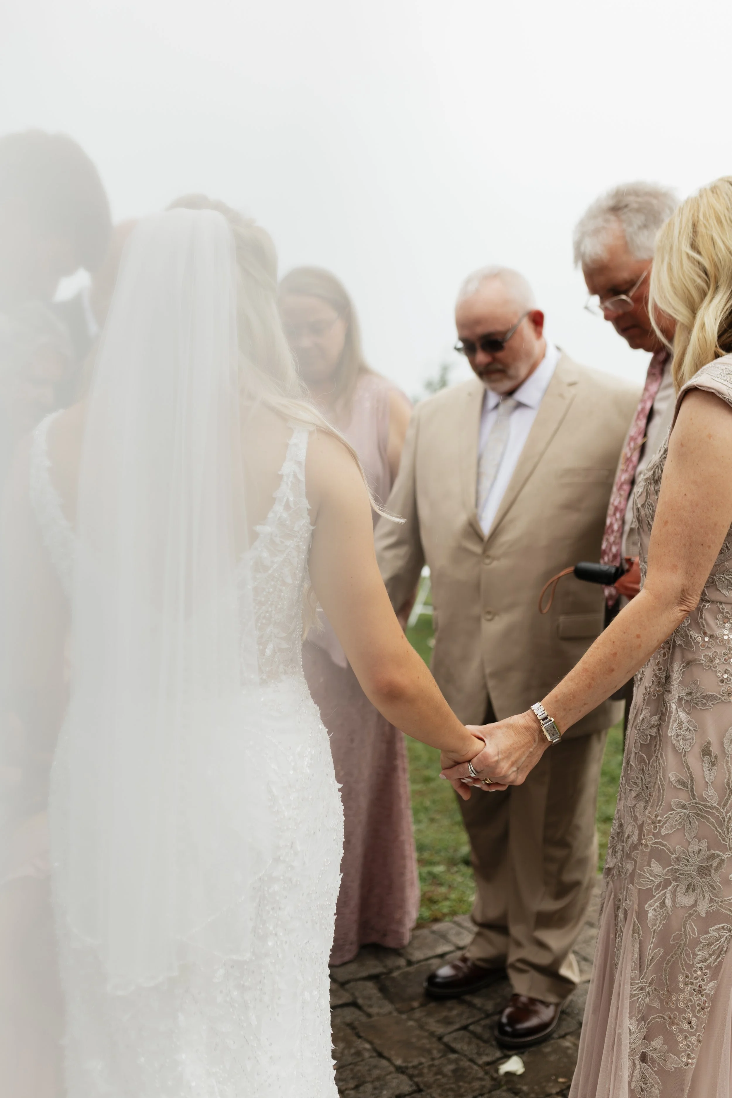 A close-up of a wedding ceremony where the bride and groom hold hands in prayer, surrounded by family members, with the focus on their clasped hands and soft-focus on the background