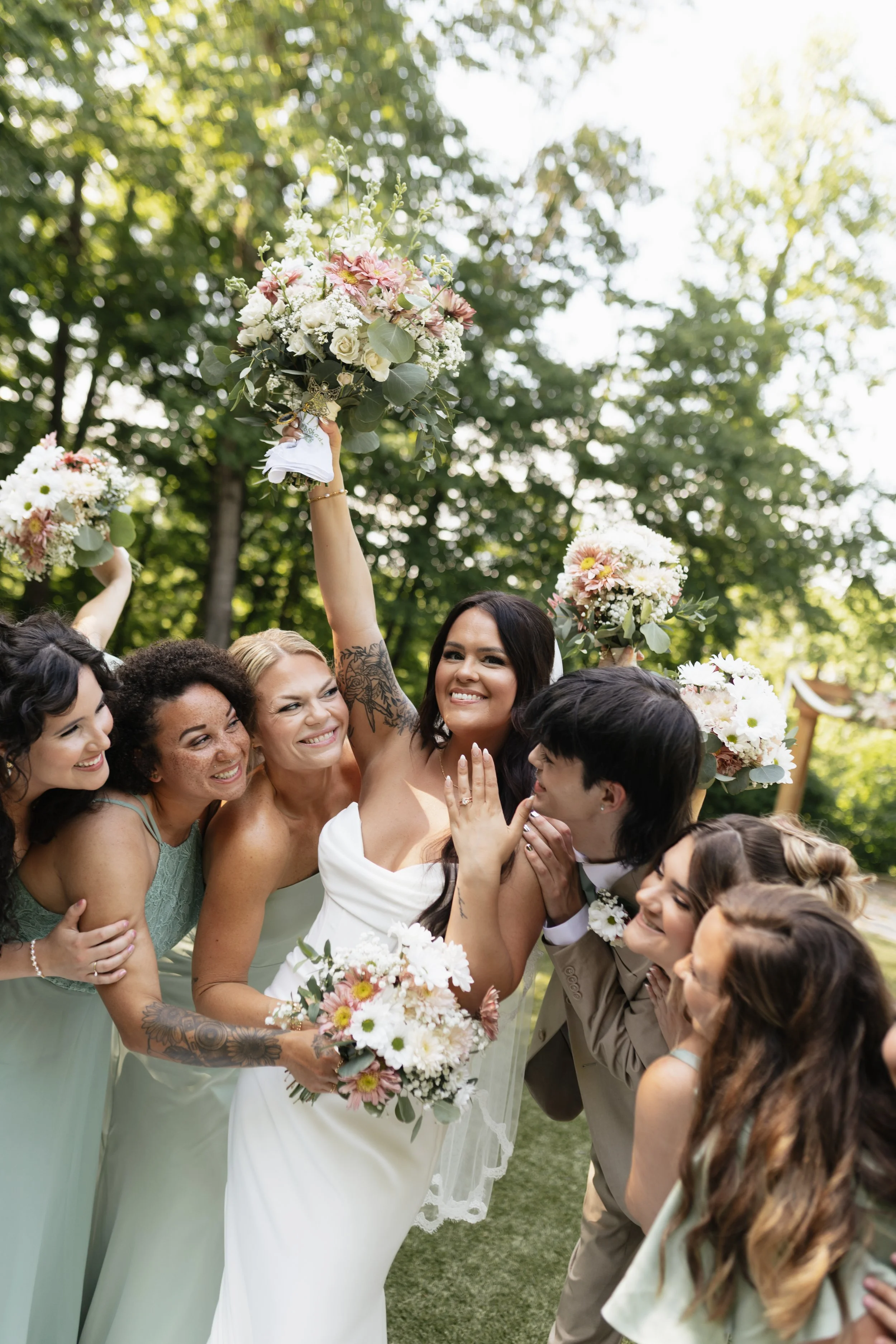 Group of women and men in wedding attire celebrating outdoors, with some holding bouquets and raising flowers in the air, surrounded by green trees.