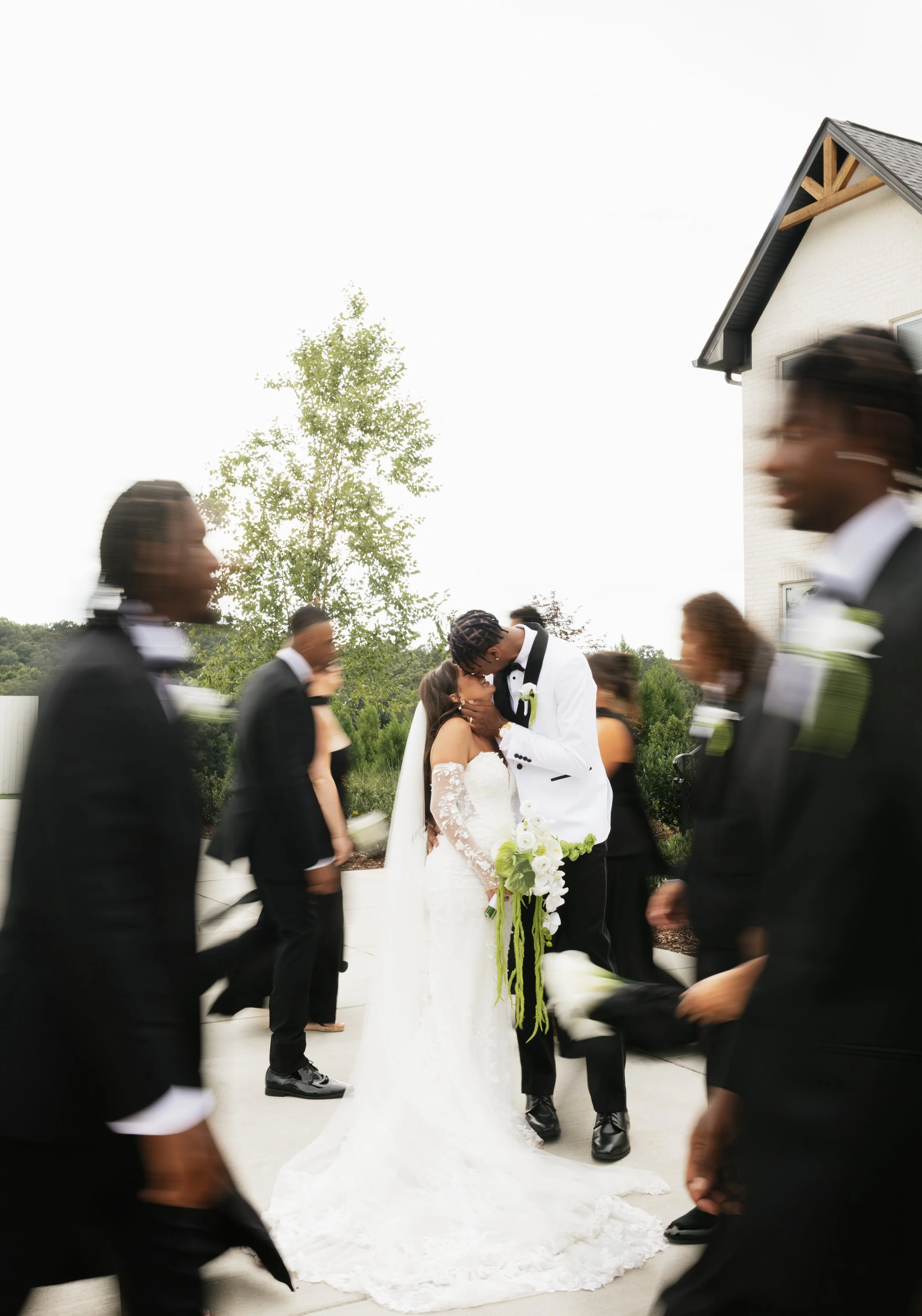 Couple sharing a kiss at their wedding surrounded by guests in black suits, outdoors with a building, trees, and a cloudy sky in the background.