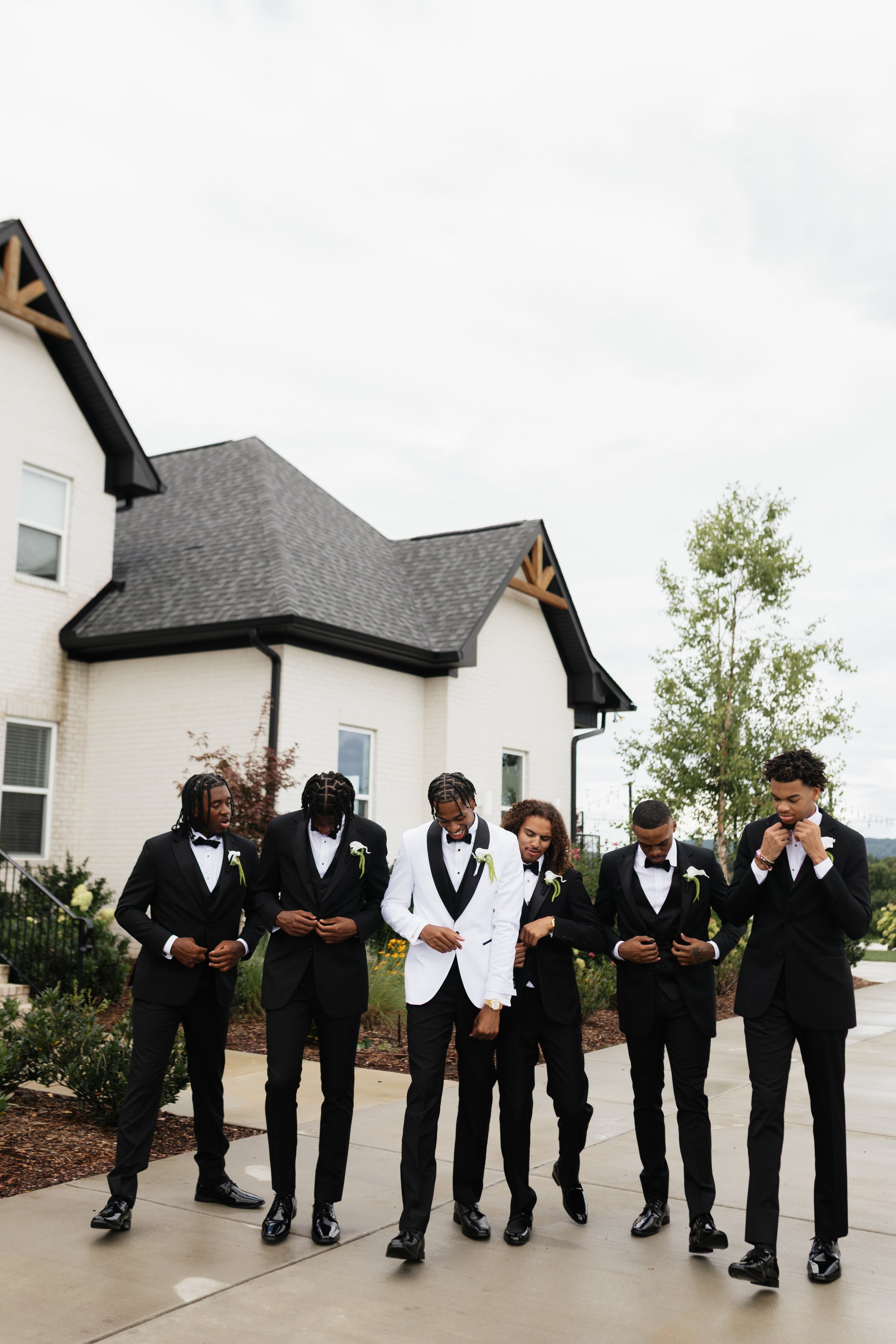 A group of men in formal tuxedos, standing in a line outdoors near a white house, with a cloudy sky above, engaged in a prayer or moment of reflection.