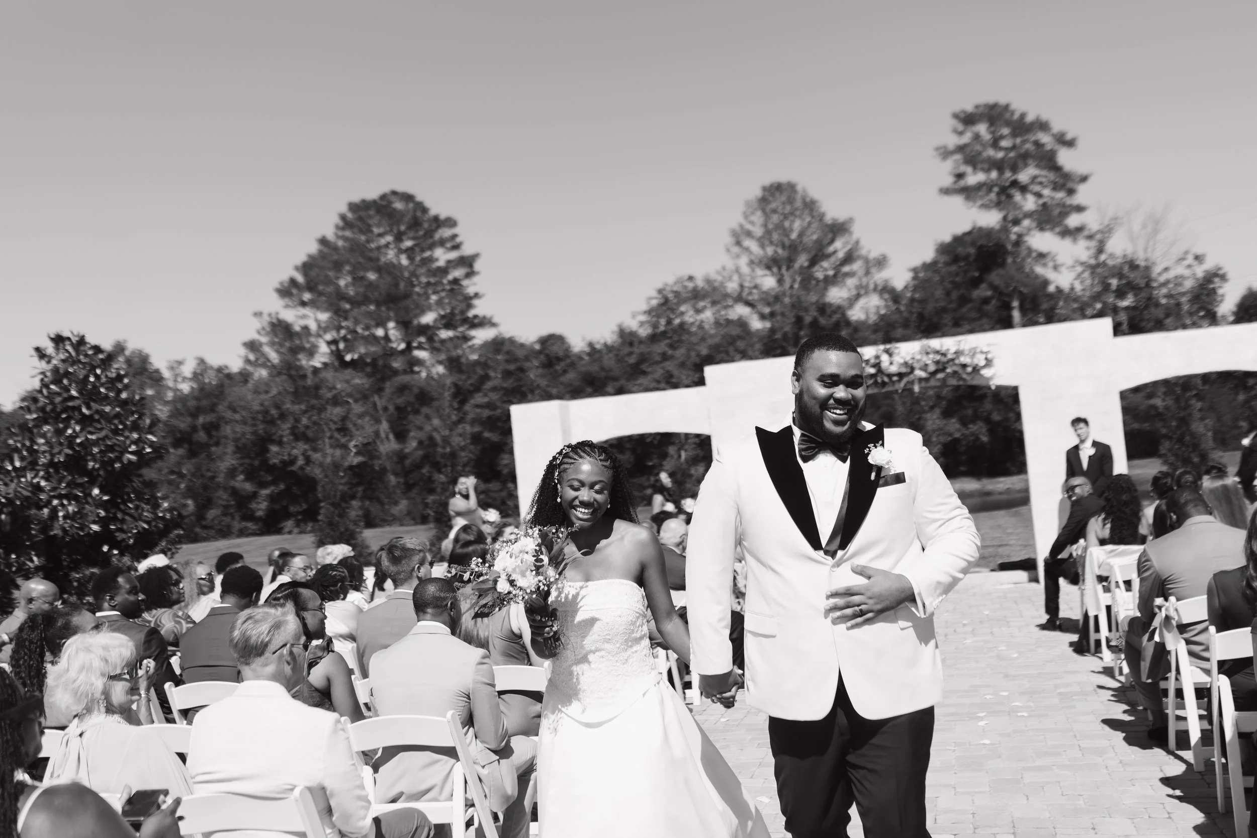 A black and white photo of a wedding ceremony outdoors with a diverse crowd. The bride and groom are smiling and holding hands; the groom is dressed in a tuxedo. Guests are seated in chairs, and trees are in the background.