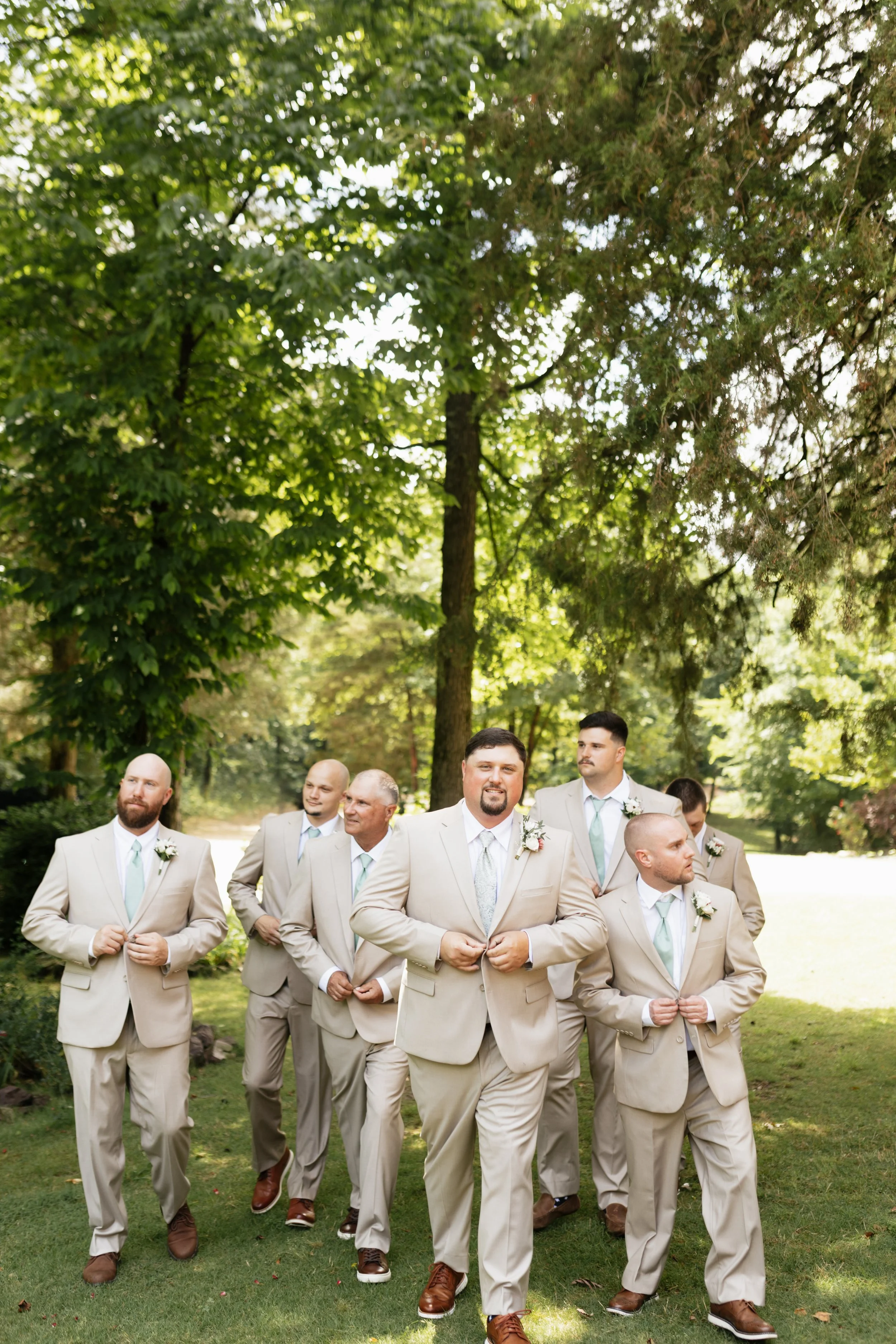 A group of men in beige suits and light blue ties walking outdoors in a park with green trees and grass, during a wedding event.