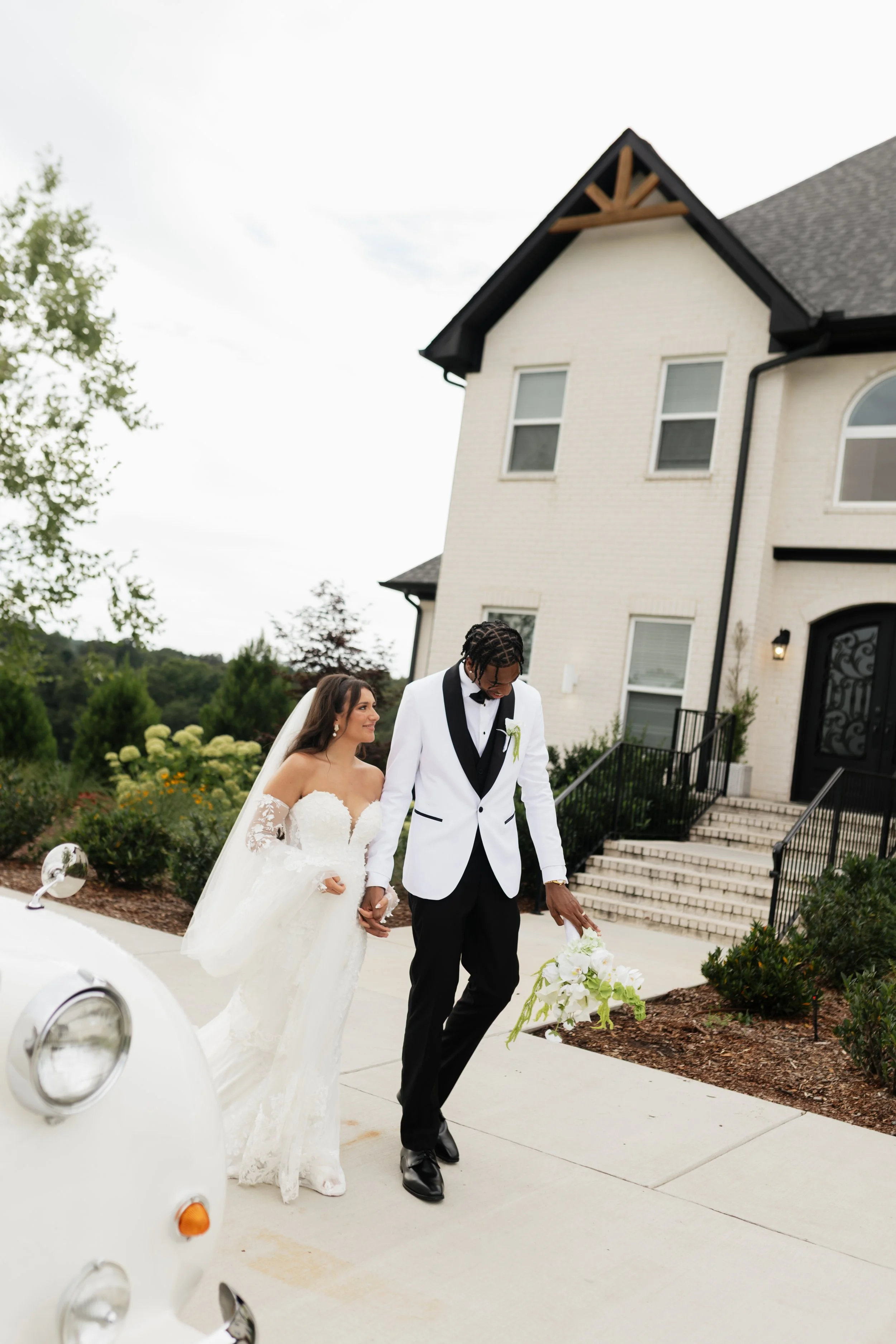 A bride and groom walking hand in hand outside a large house during daytime, with the bride in a white wedding gown and the groom in a white tuxedo jacket and black pants, holding a bouquet of white flowers.