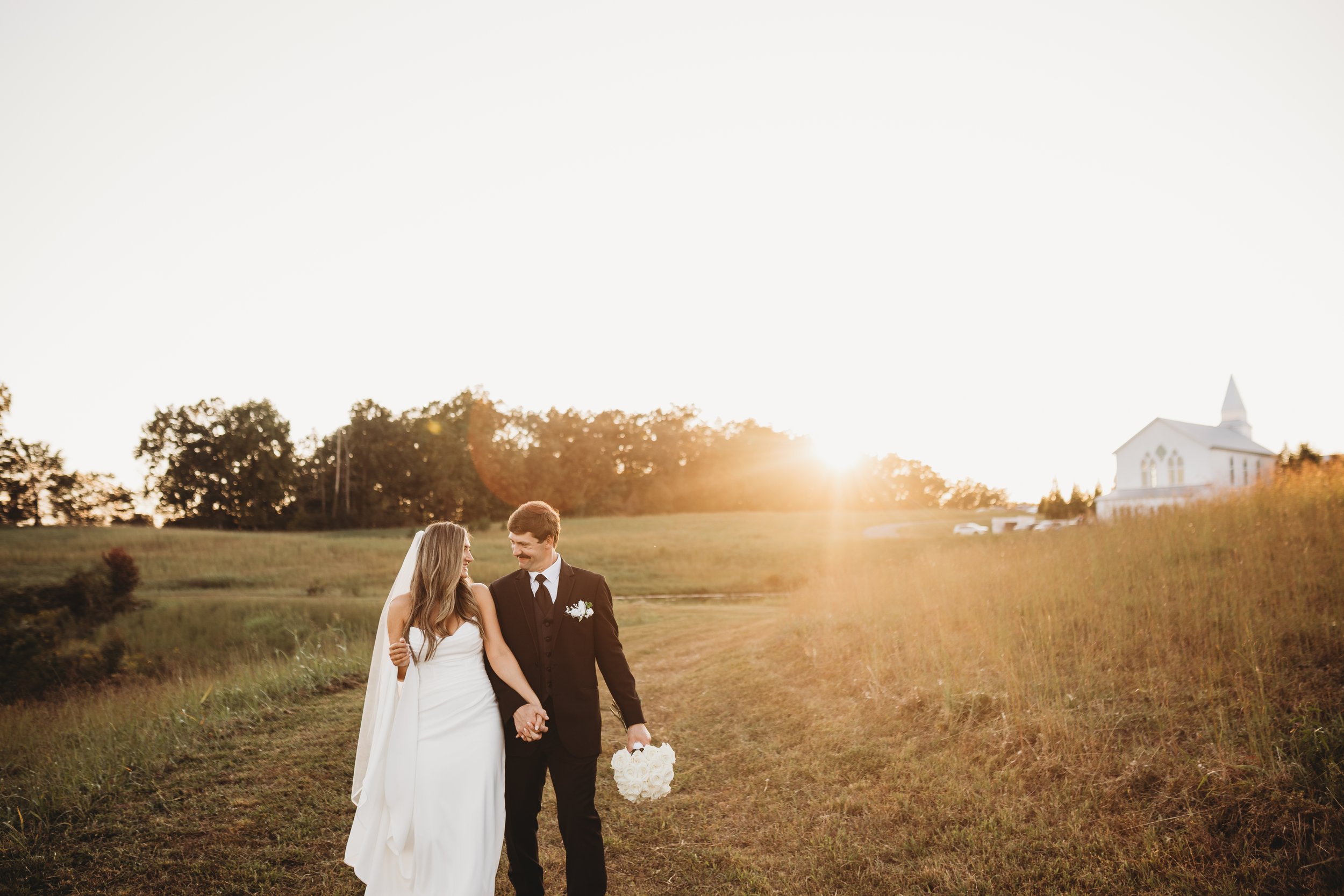 A bride and groom walking hand in hand outdoors during sunset, with the bride holding a bouquet of white flowers and gazing at the groom. The background features a church on a hill, trees, and a wide open grassy field.