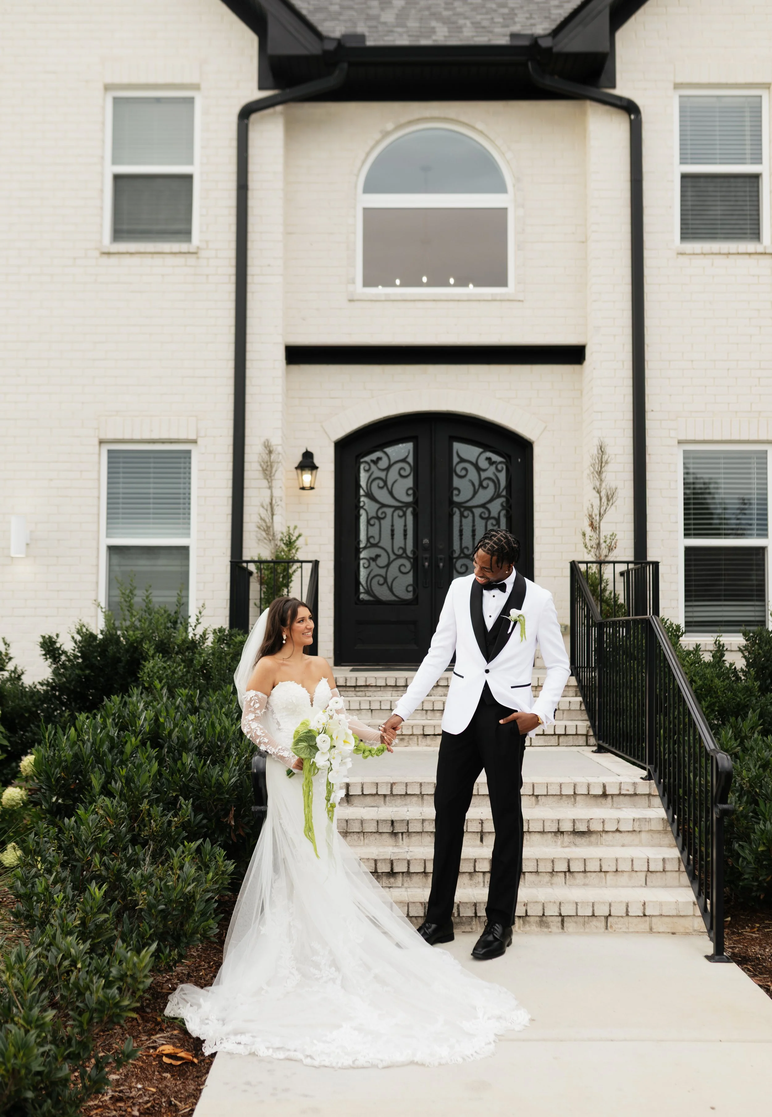 A bride and groom holding hands on the steps of a large, white brick house with black front door and windows, with greenery on either side, during daytime.