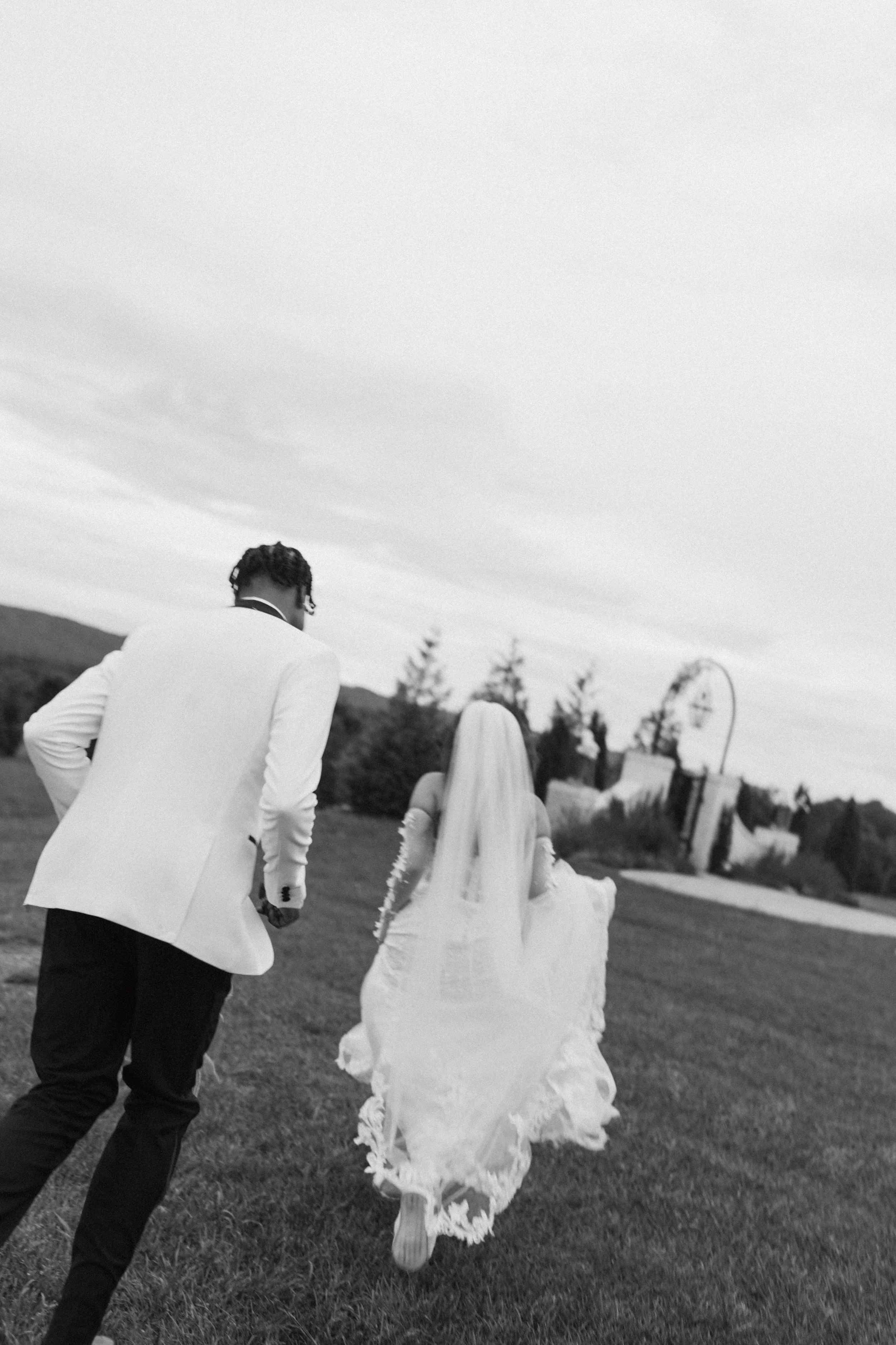 A black-and-white photograph of a couple holding hands and walking outside. The woman is wearing a long dress and a headscarf, and the man is wearing a white jacket and dark pants. They are walking on a lawn with trees and outdoor structures in the b