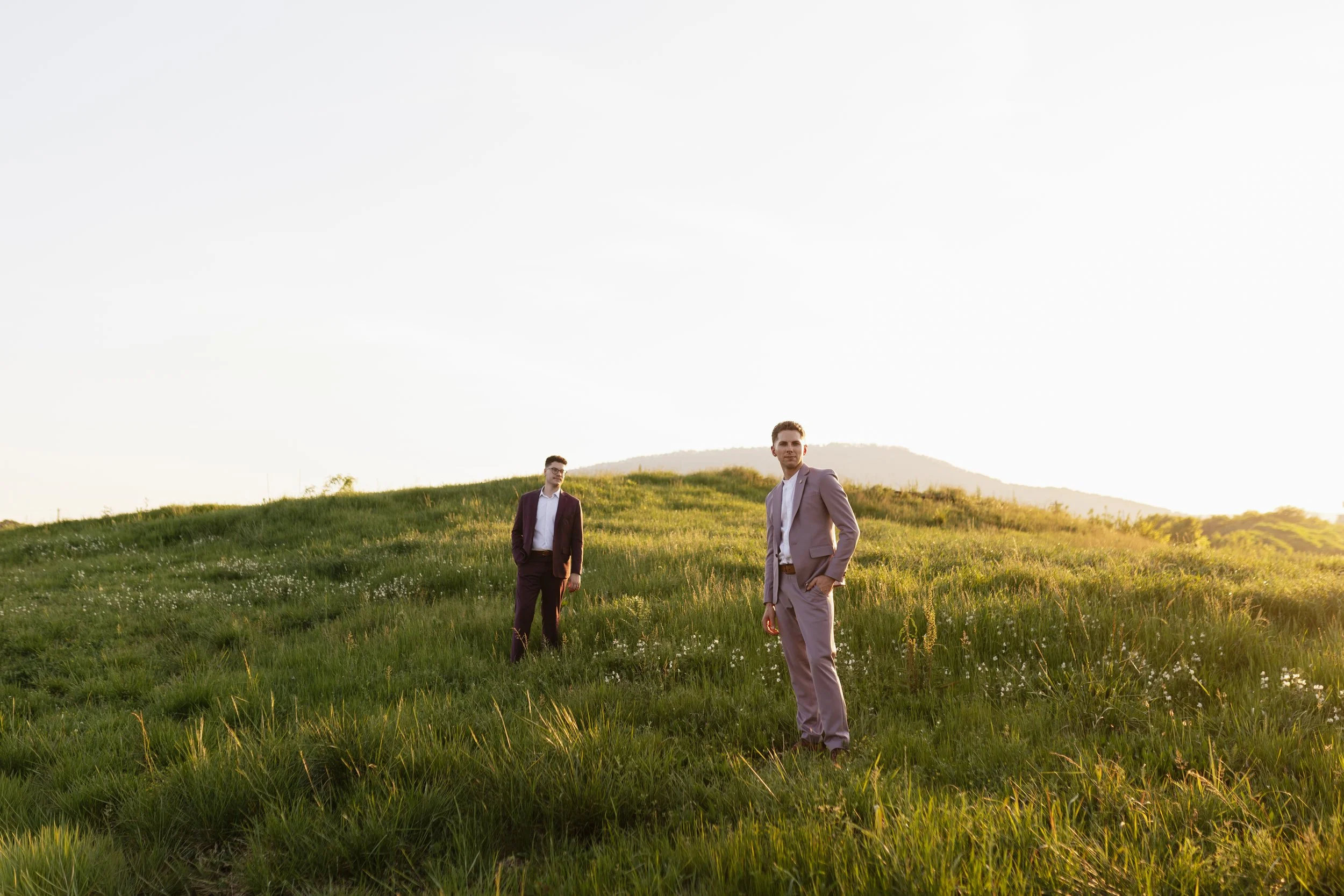 Two men in suits standing on a grassy hill at sunset.