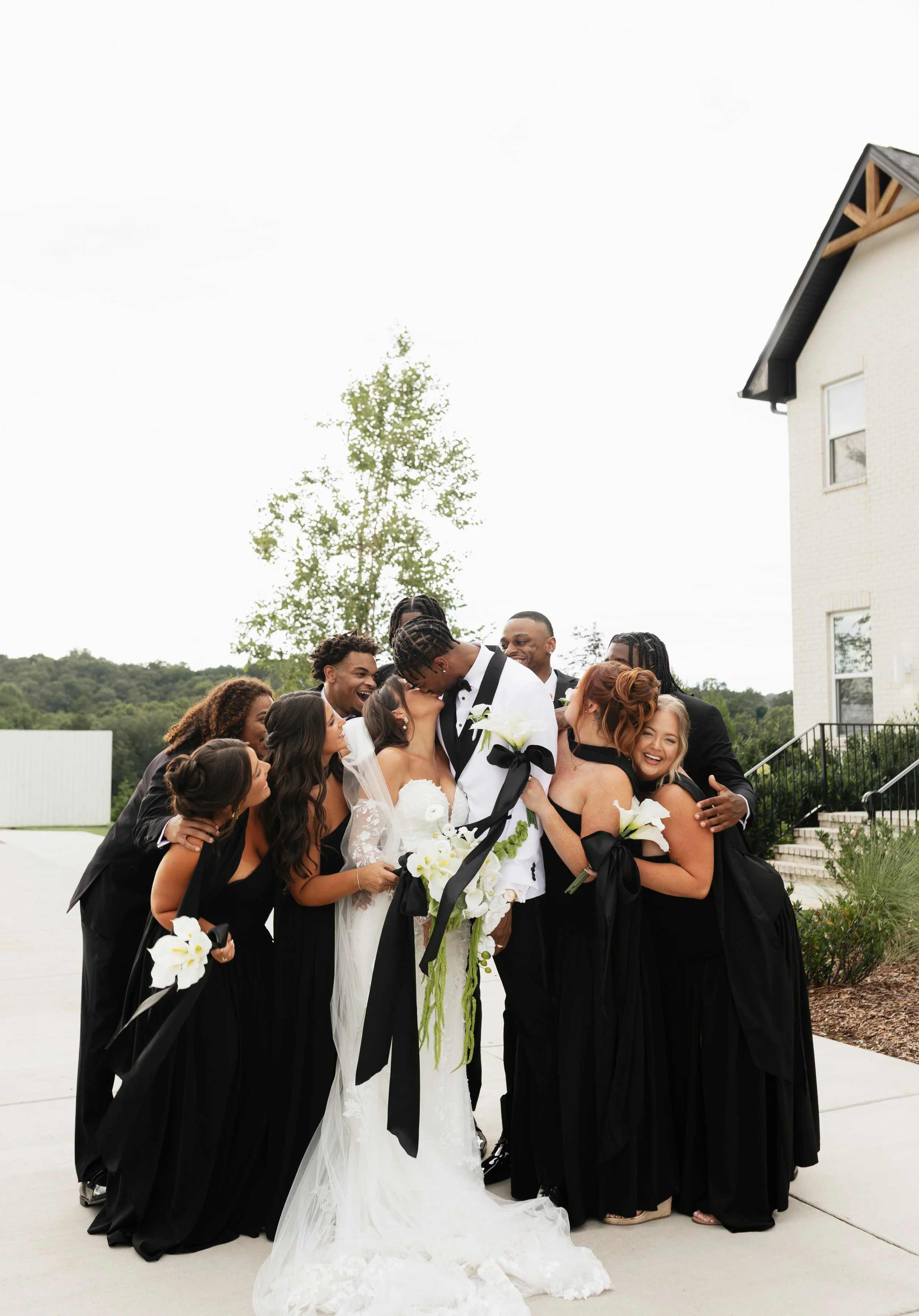 A wedding party consisting of the bride, groom, and several bridesmaids and groomsmen smiling and hugging outdoors near a white building and greenery.