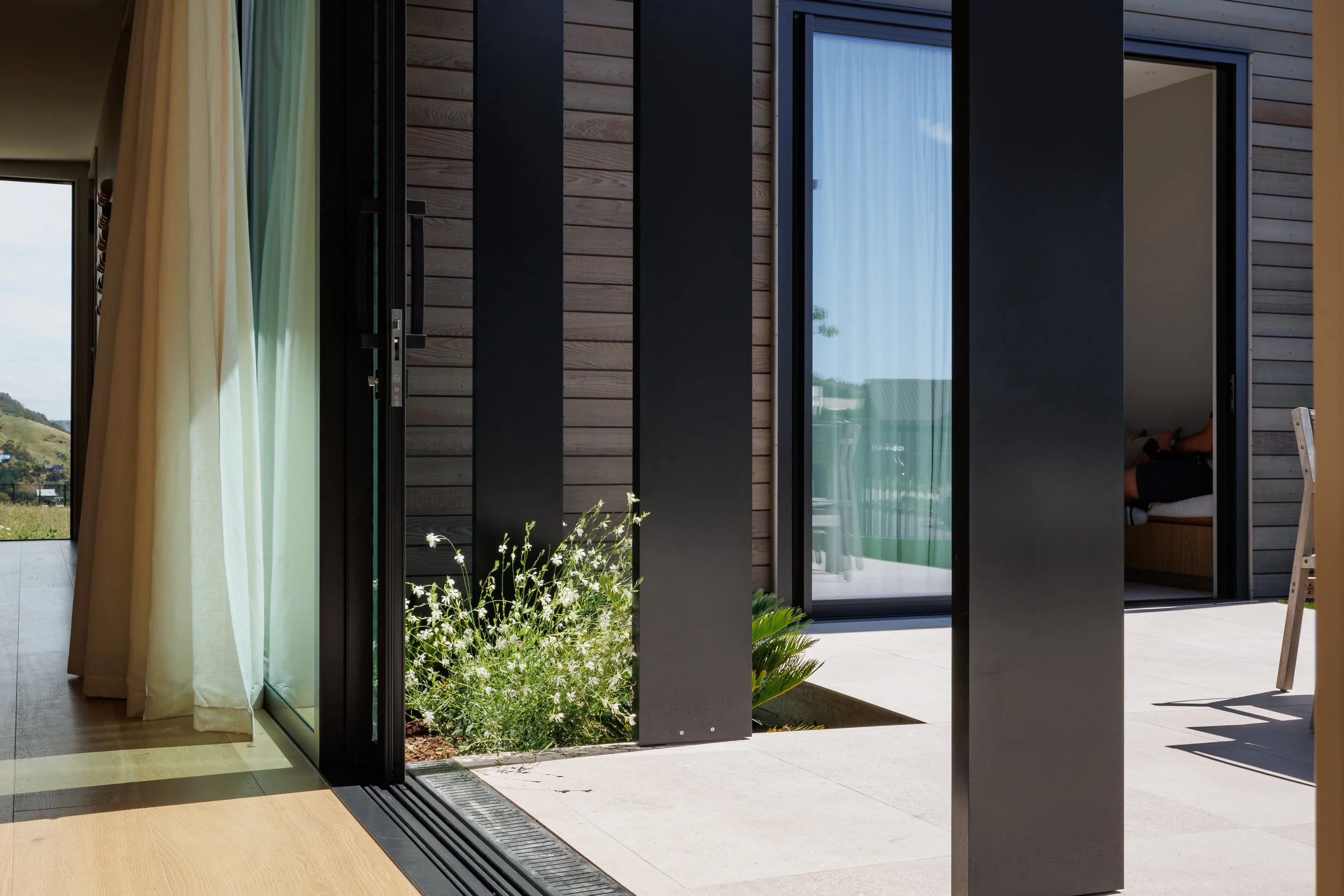 Modern sliding glass doors opening to an outdoor patio with hills in the background, inside there are curtains and a glimpse of a person sitting on a sofa.