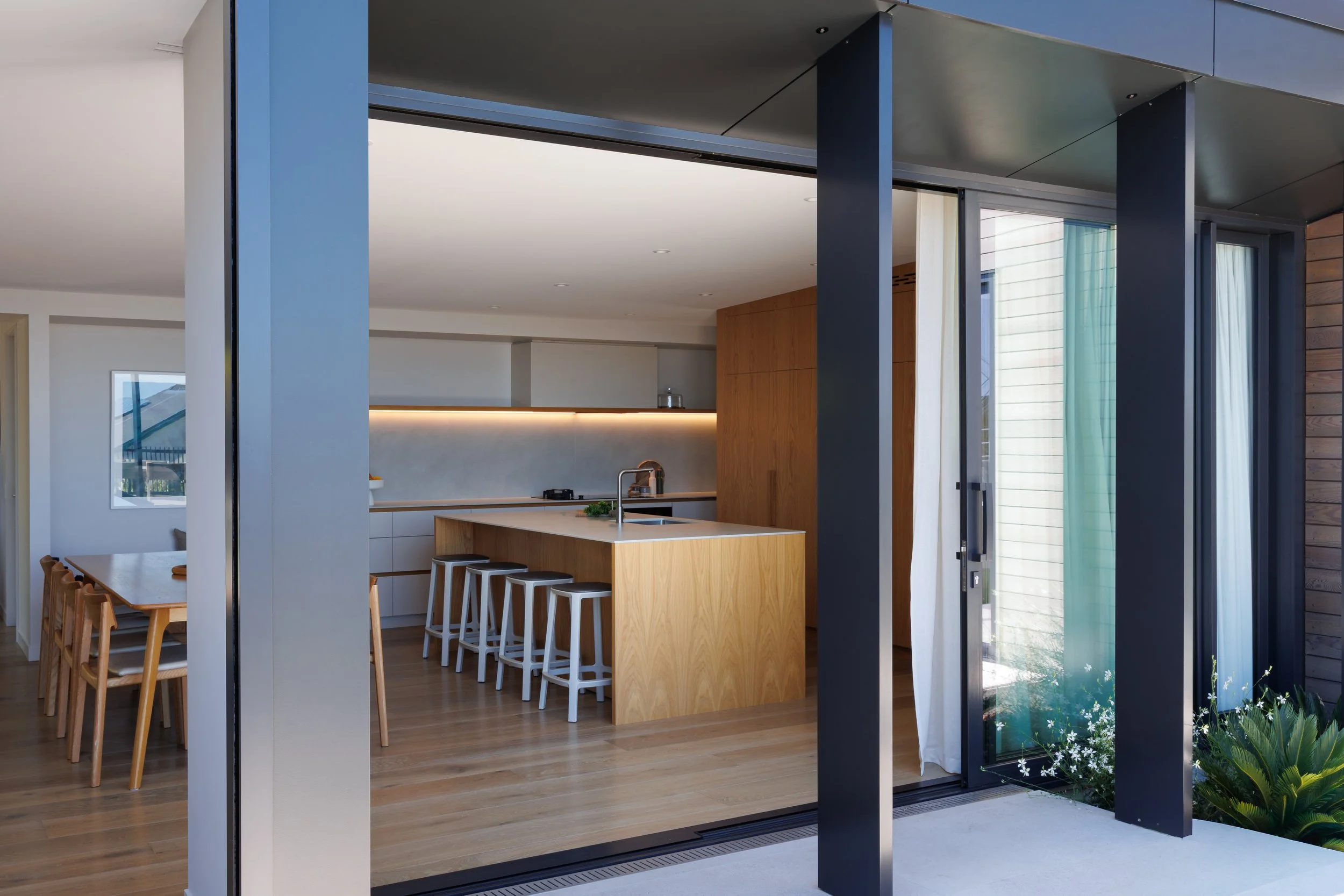 Modern kitchen with a wooden island, bar stools, sliding glass doors with curtains, and interior dining area with a wooden table and chairs.
