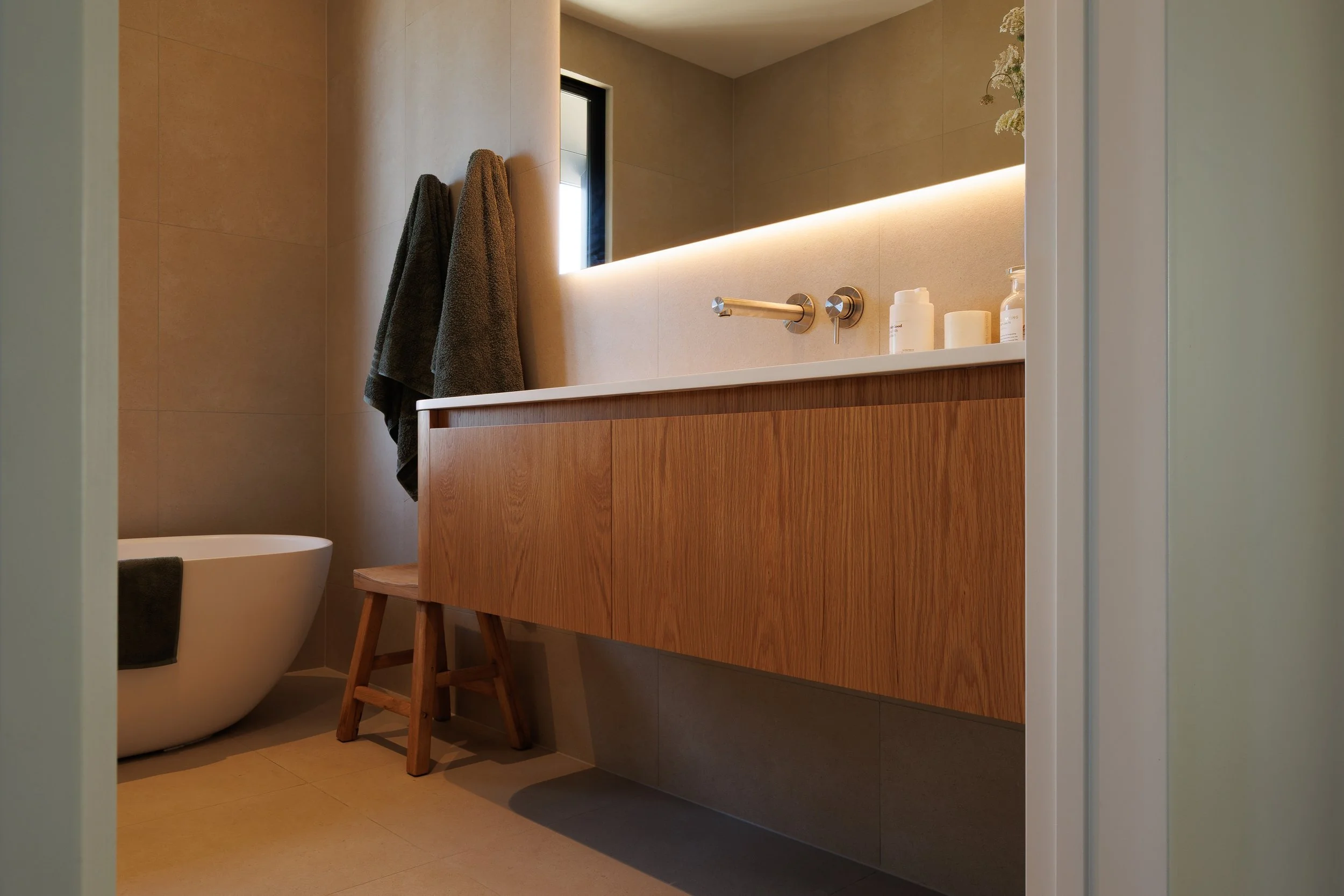 A modern bathroom with a floating wooden vanity, a large backlit mirror, towels hanging on the wall, and a partial view of a toilet.