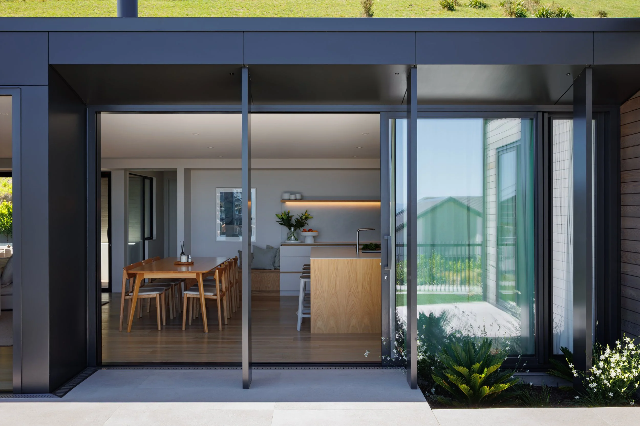 Modern house with large glass sliding doors revealing a dining area with a wooden table and chairs inside, greenery outside, and a small garden with plants and flowers.