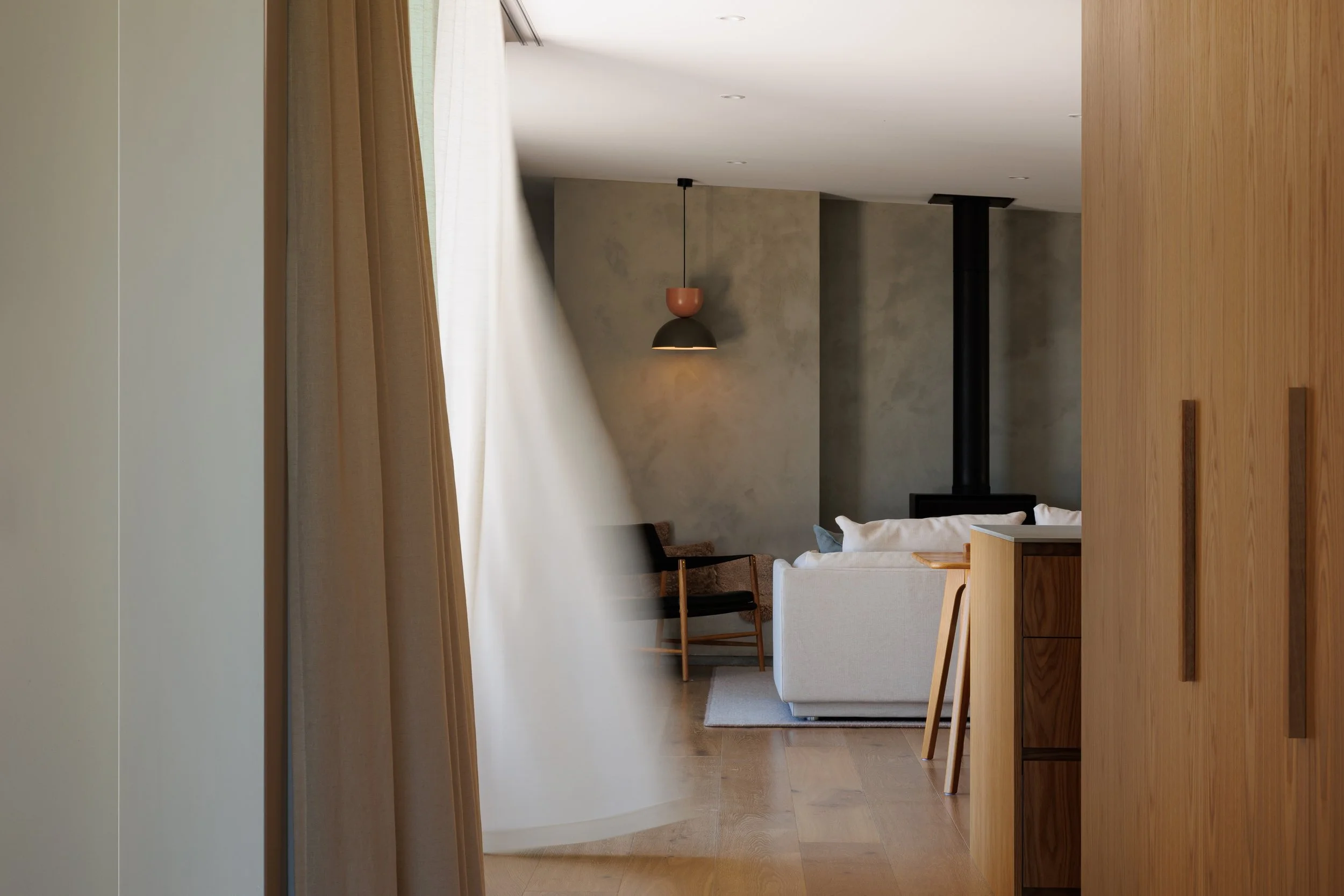 A modern, minimalistic living room with a white sofa, wooden chairs, and a textured concrete wall. A black and terracotta pendant light hangs from the ceiling, and natural light filters through beige curtains.