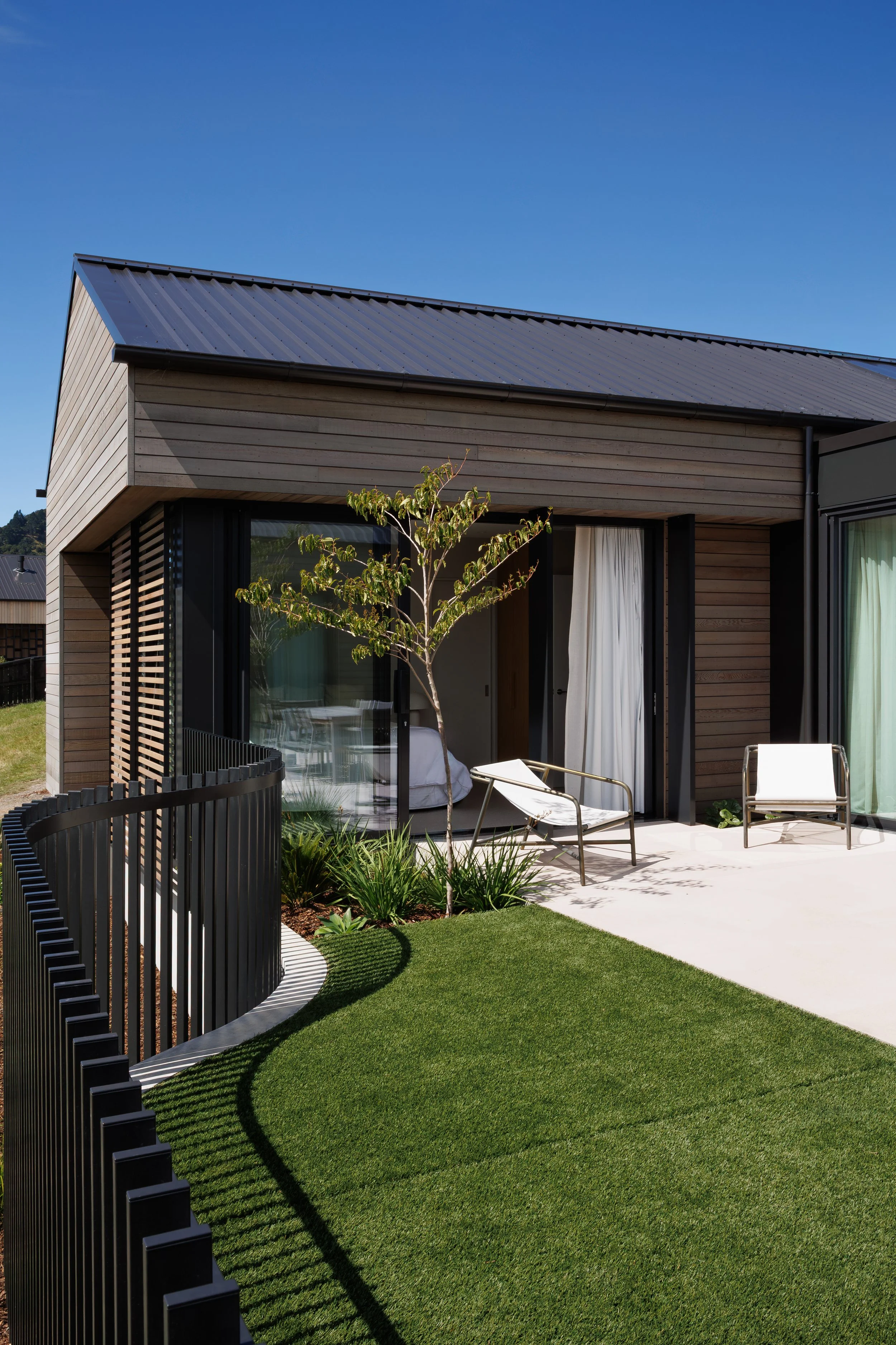 Modern house with wooden exterior, glass doors, a small tree, outdoor chairs, and a well-maintained lawn under a clear blue sky.