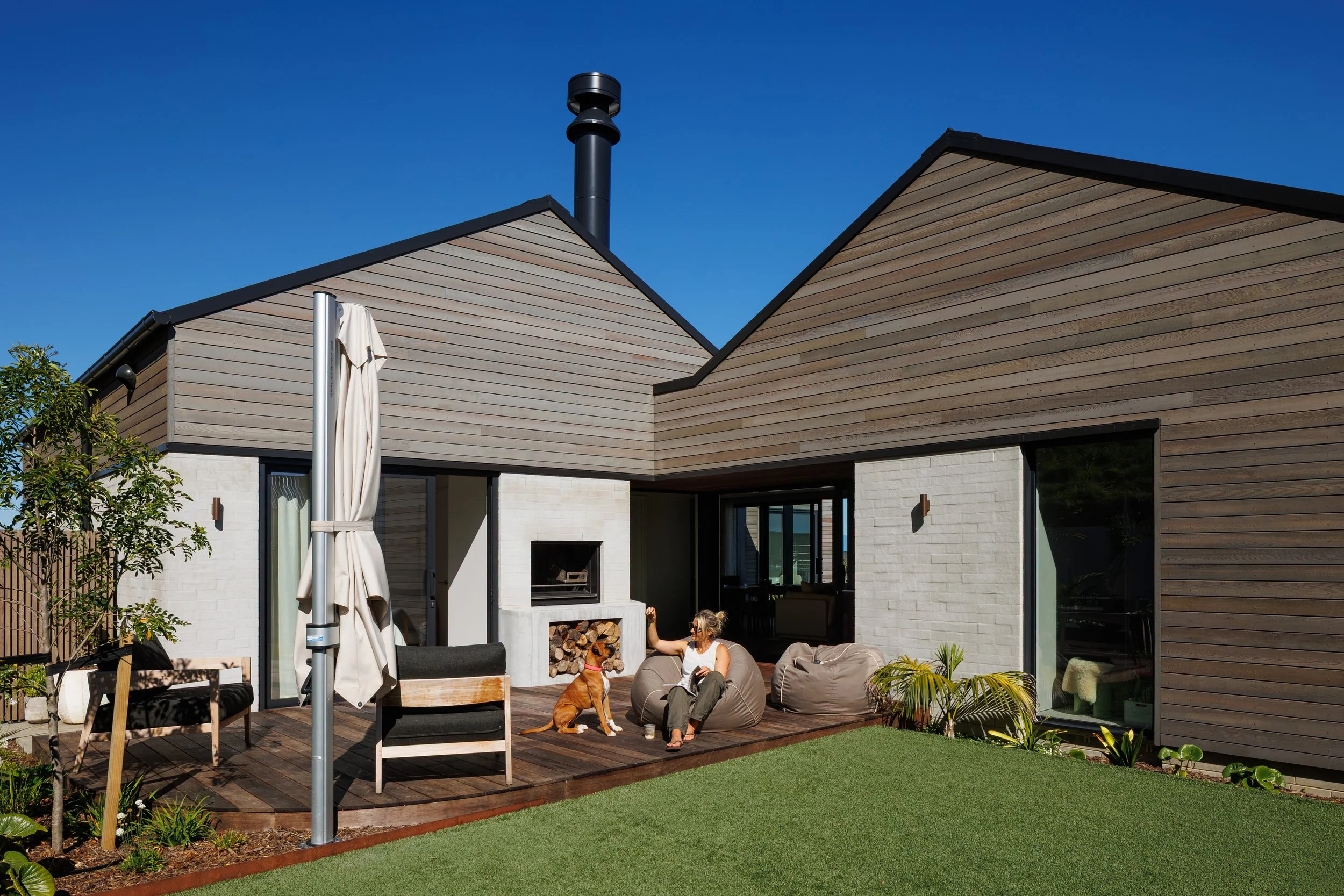 Modern house with wooden siding and a black chimney, a woman sitting on outdoor furniture with a dog nearby, and a backyard deck with plants and a small grassy area.