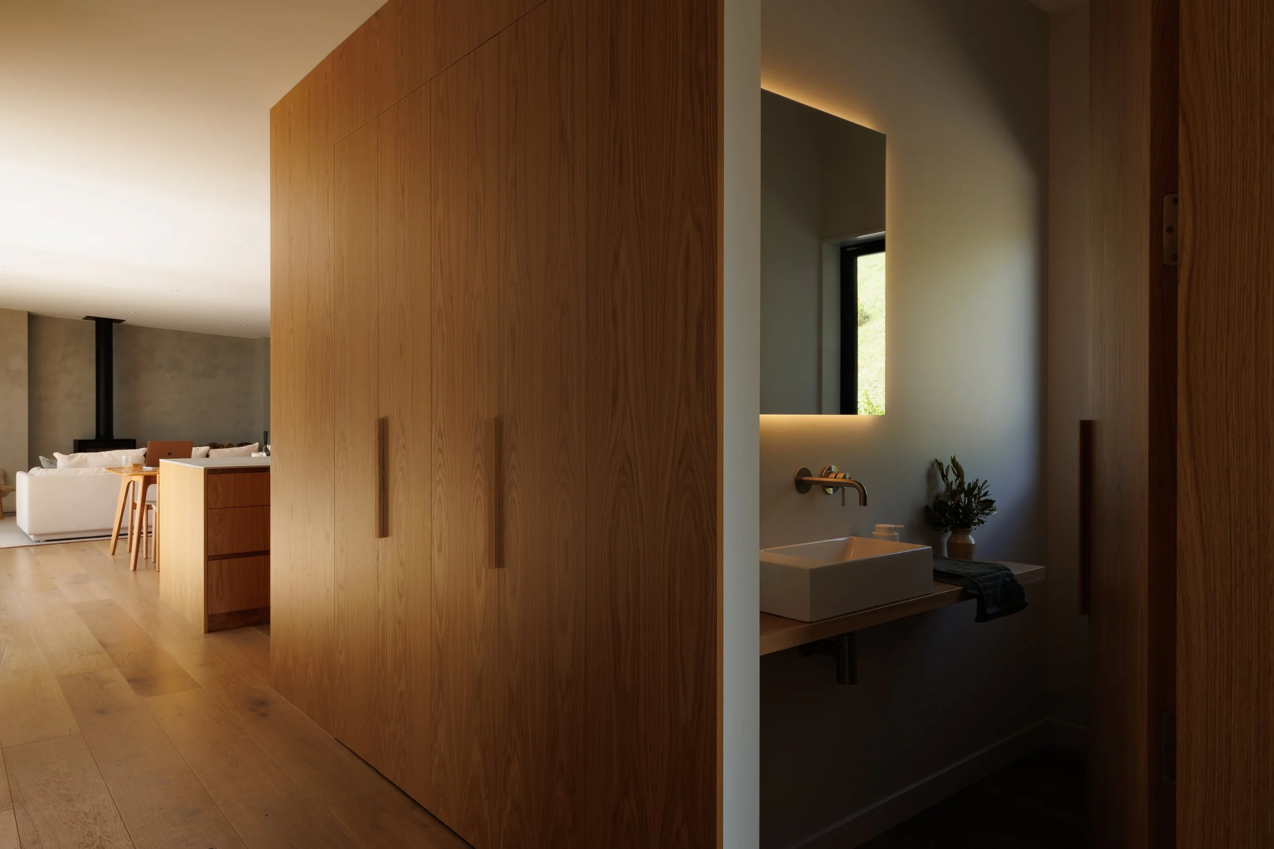Interior view of a modern home with wooden walls, a glimpse of a living room with a white couch, and a bathroom with a white sink, a black faucet, and a window with greenery outside.