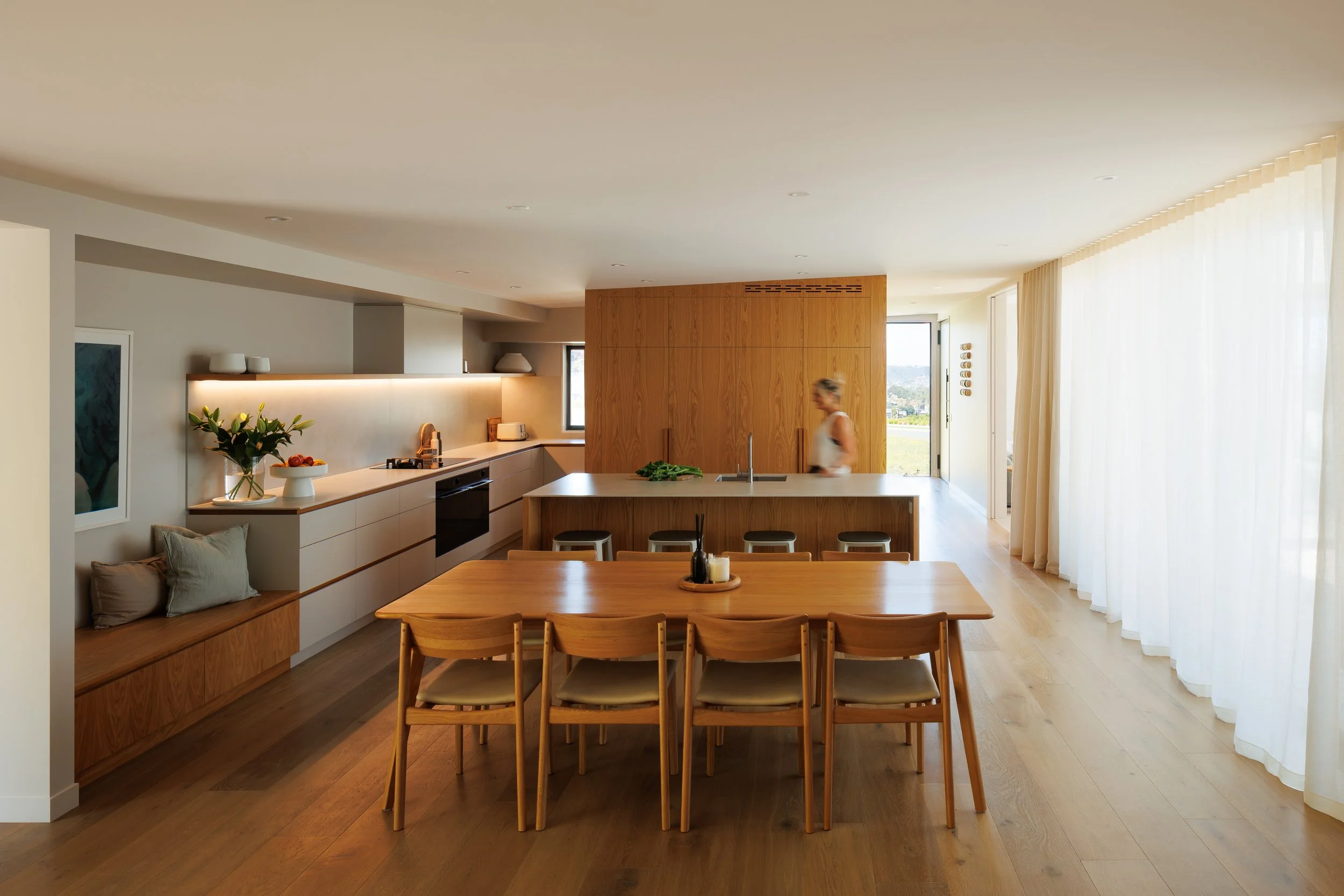 A modern, minimalist kitchen and dining area with wooden furniture, a large wooden cupboard, white cabinetry, a vase with flowers, and large windows with sheer curtains, illuminated by natural light.